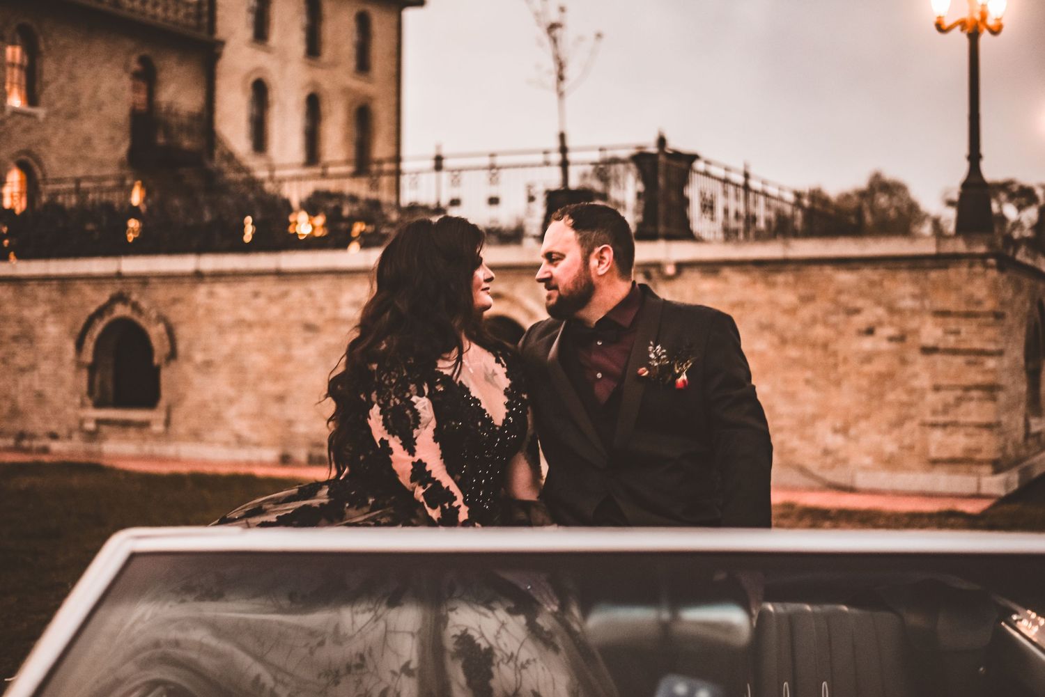 Romantic vintage-style photo with couple by classic car at dusk near stone building.