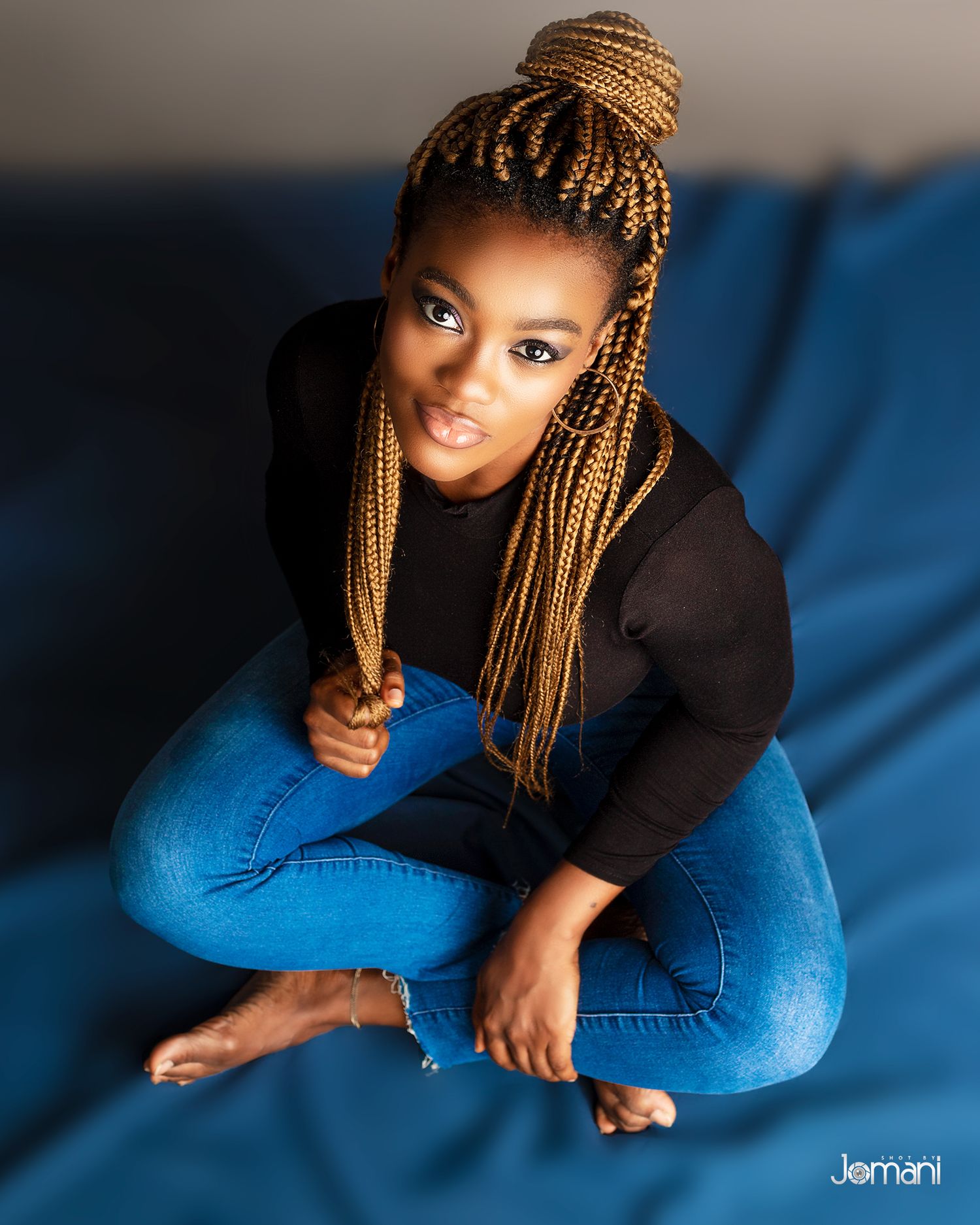 Young woman with golden box braids poses cross-legged on blue surface wearing black top and jeans.