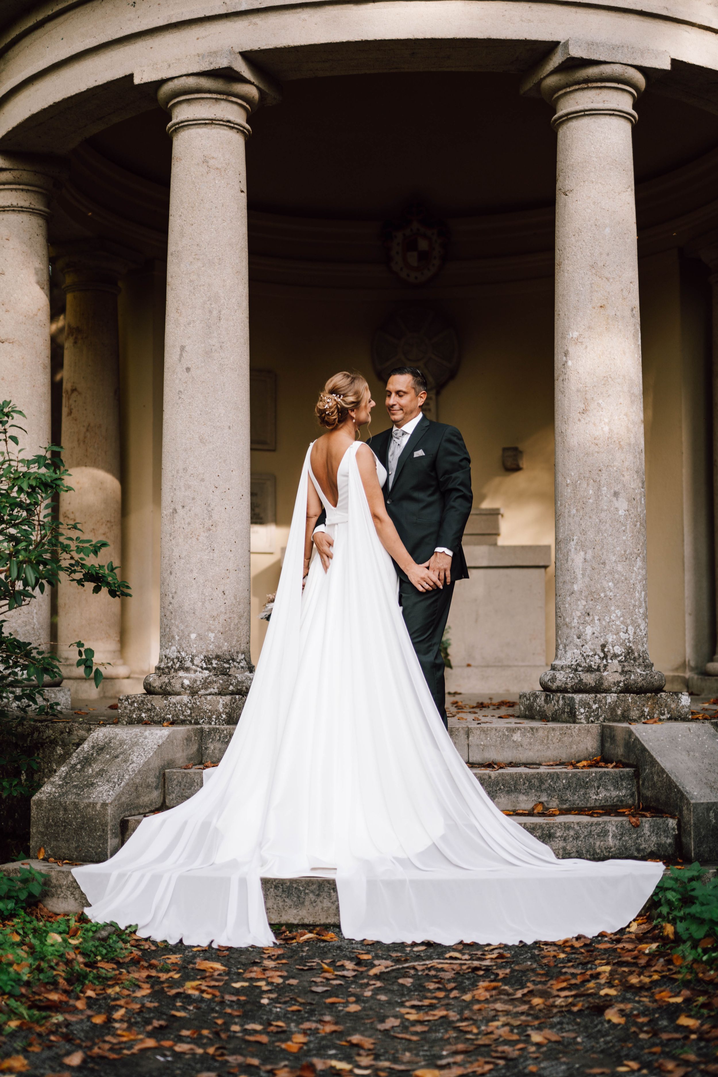 Elegant wedding couple poses against stone columns with autumn leaves scattered on the ground.