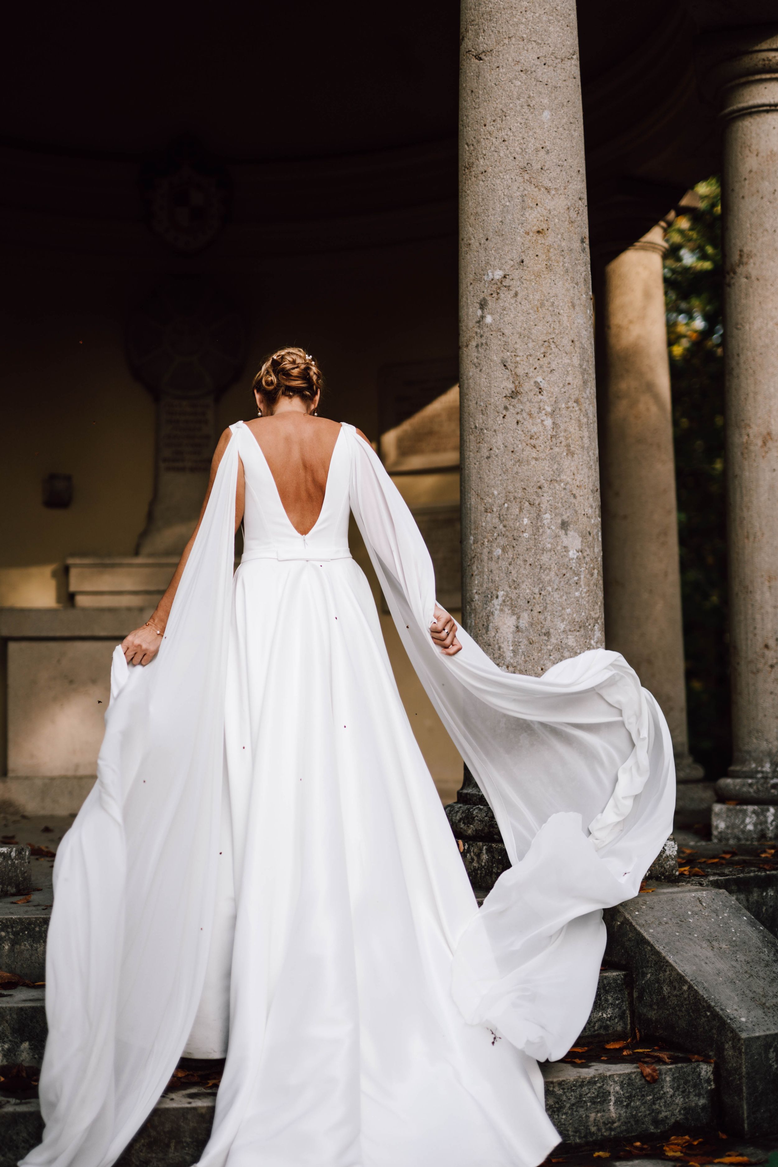 Ethereal wedding dress with dramatic cape photographed against stone colonnade.