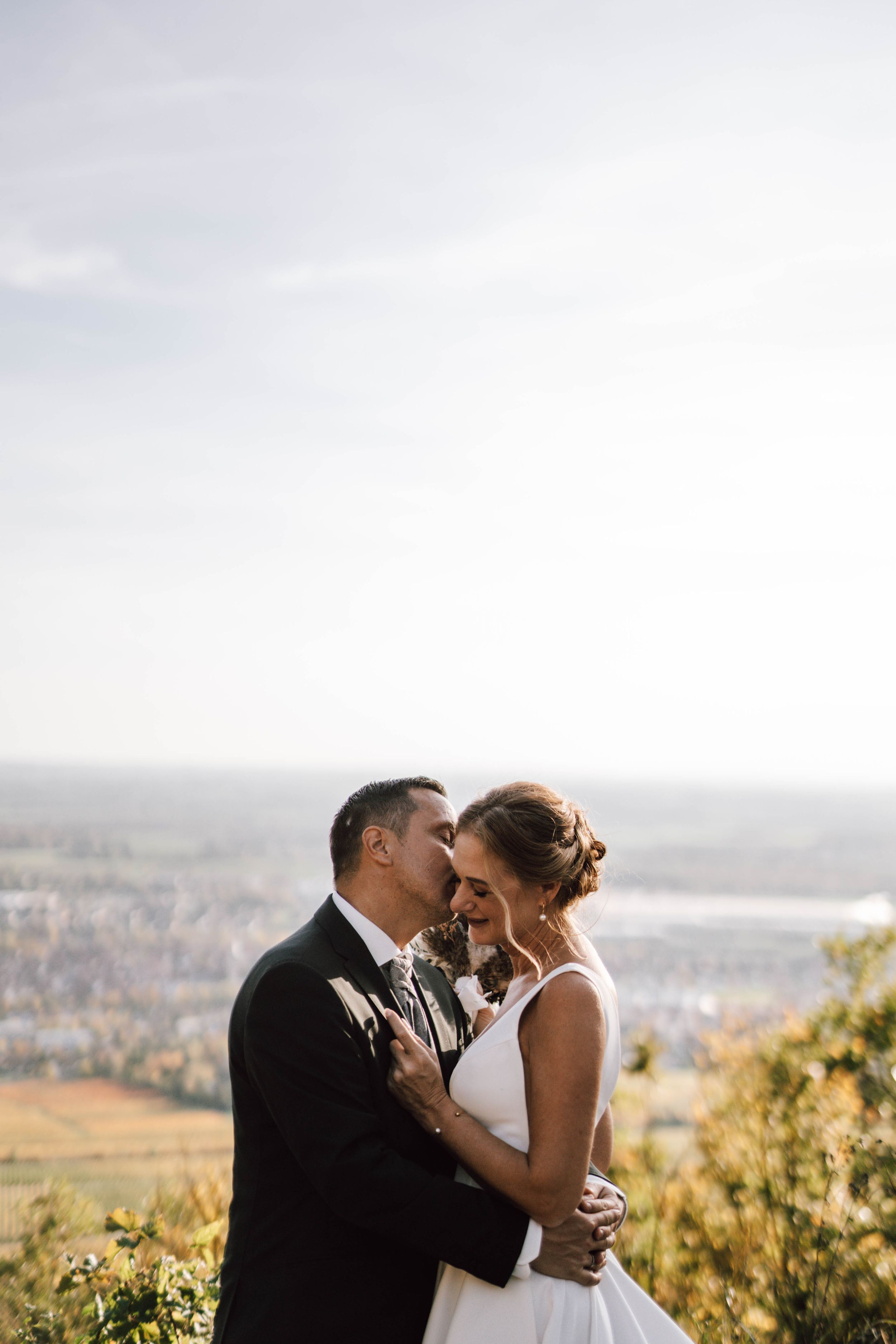 Romantic wedding couple shares an intimate moment on a scenic hilltop overlooking a valley at sunset.
