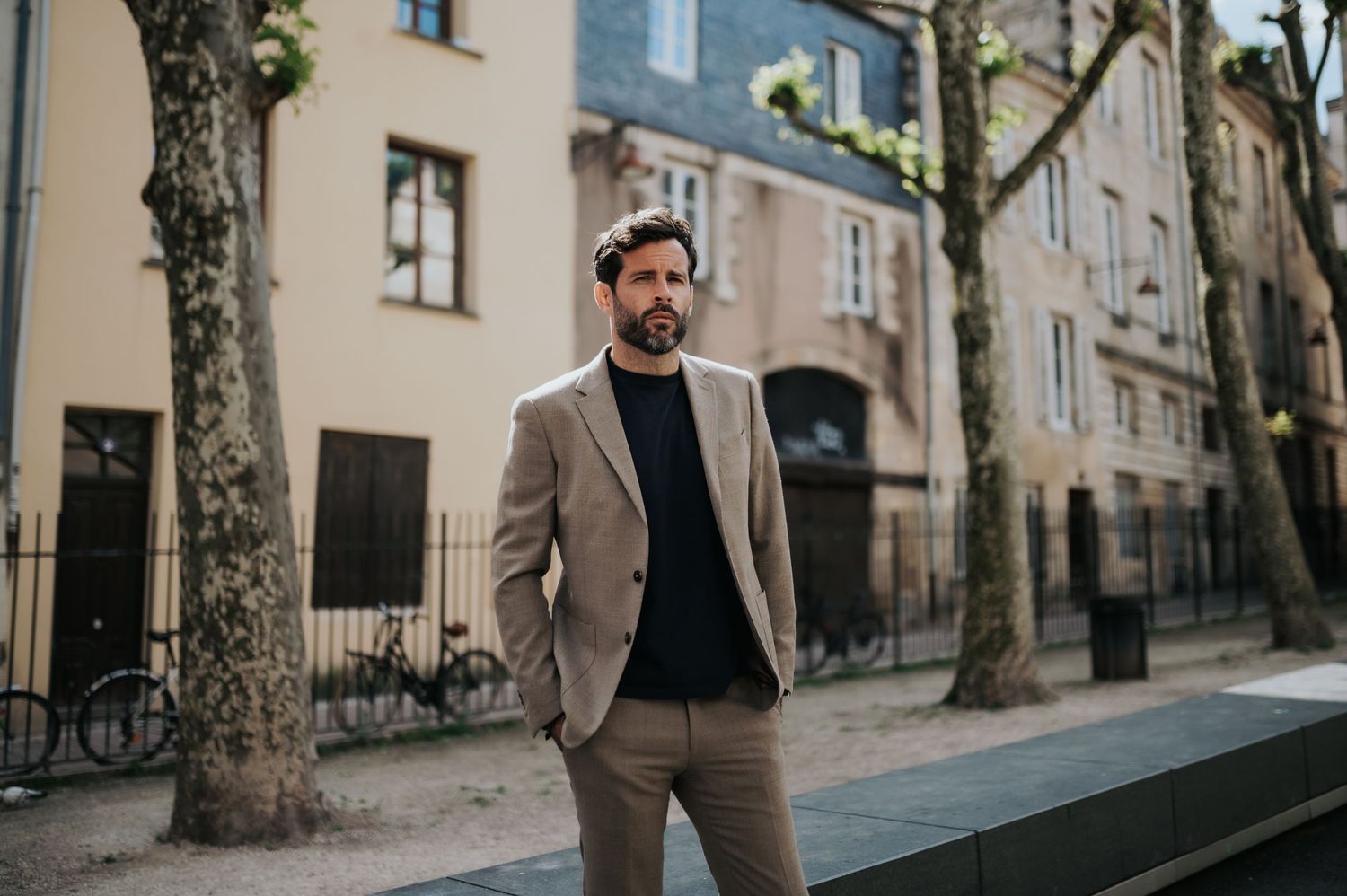Man wearing tan suit and dark sweater poses on tree-lined urban street.