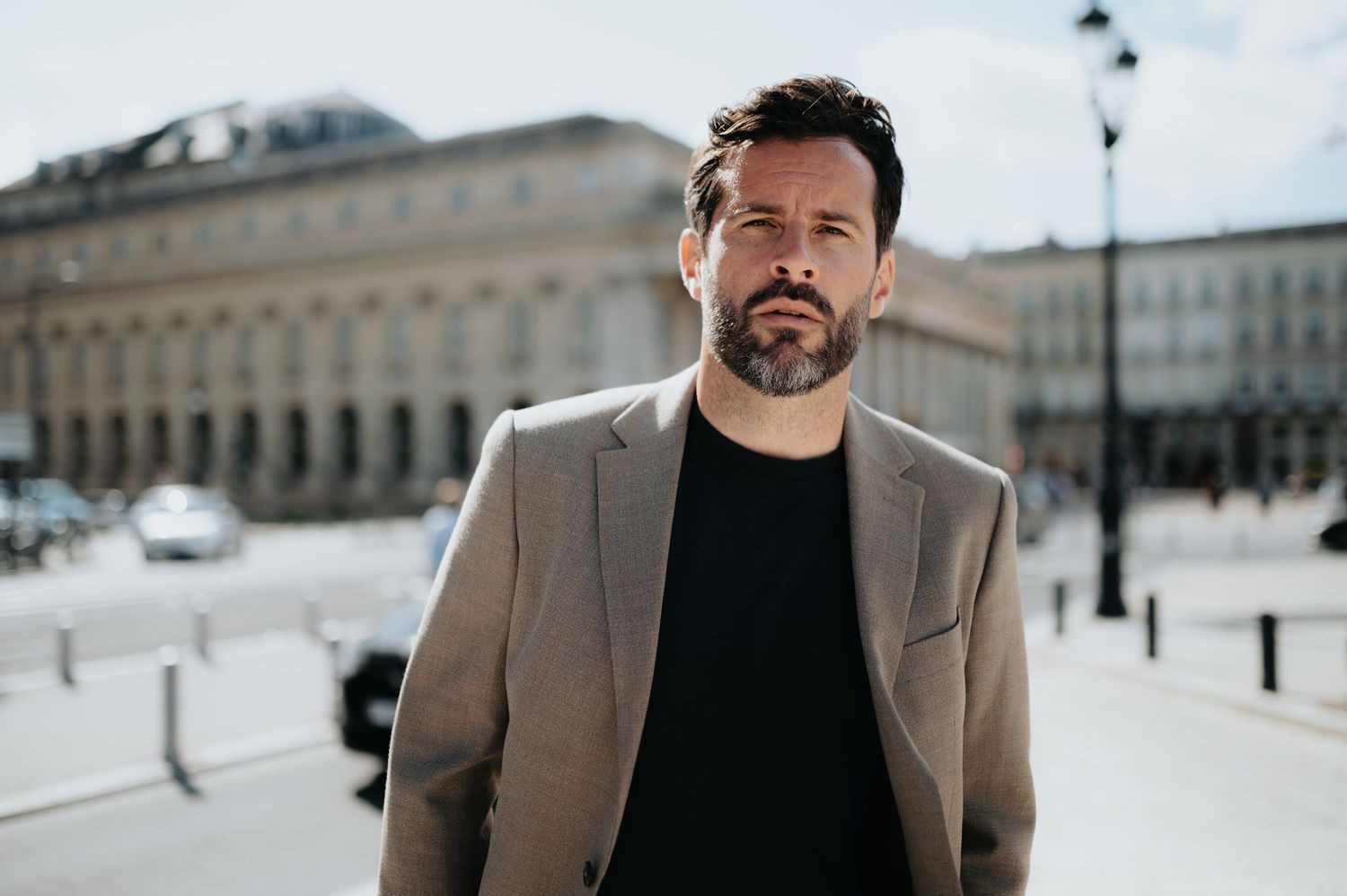 Man in beige blazer and black shirt walking through a European city square.