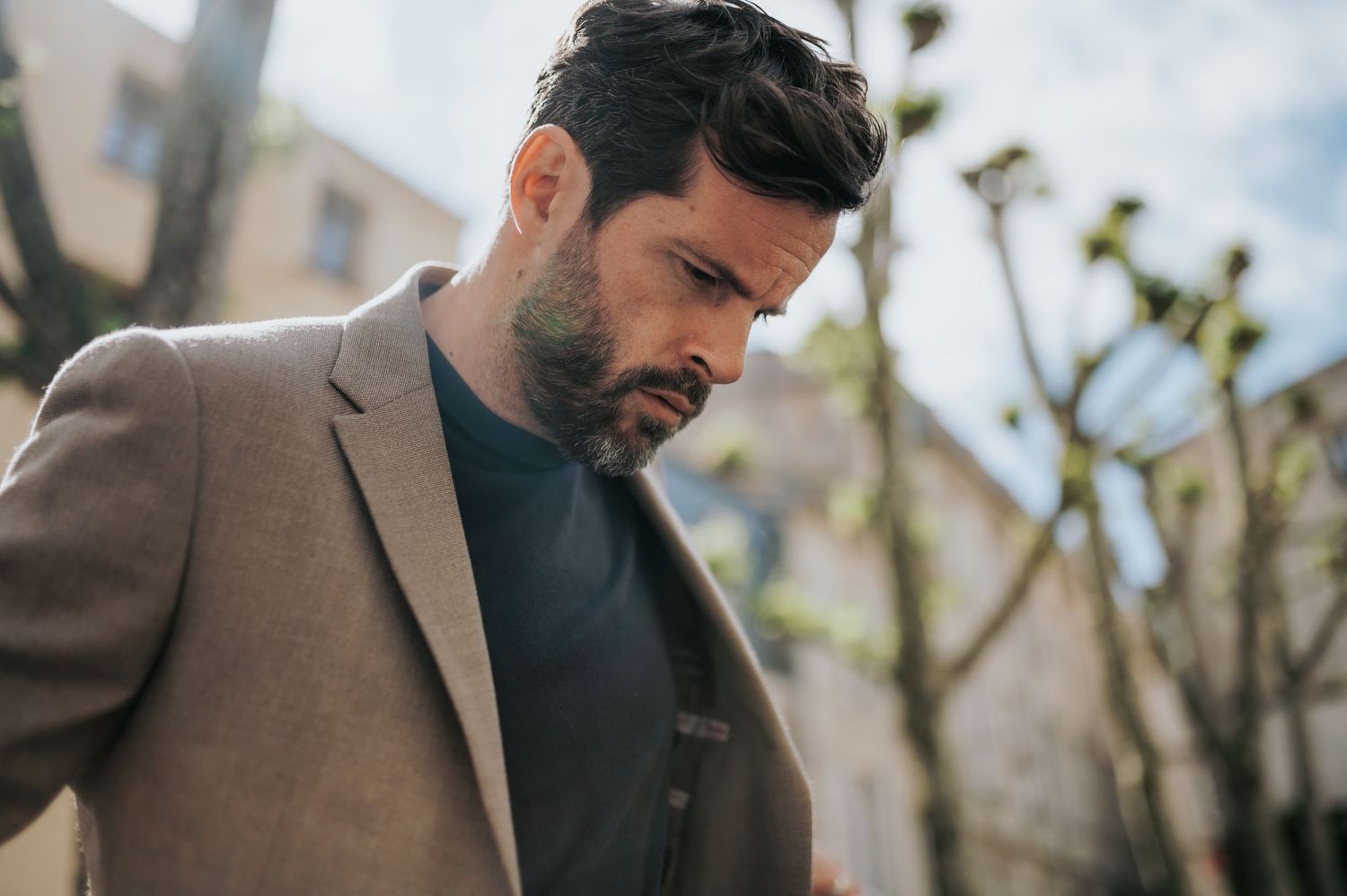 Close-up profile shot of man in beige blazer and dark sweater against blurred background.