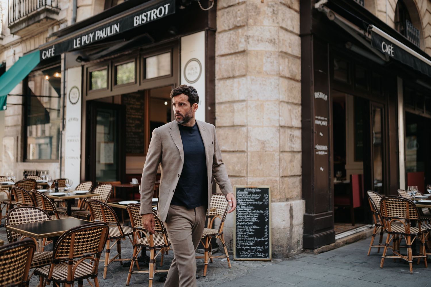 Stylish man in blazer and turtleneck standing outside Parisian cafe with outdoor seating.