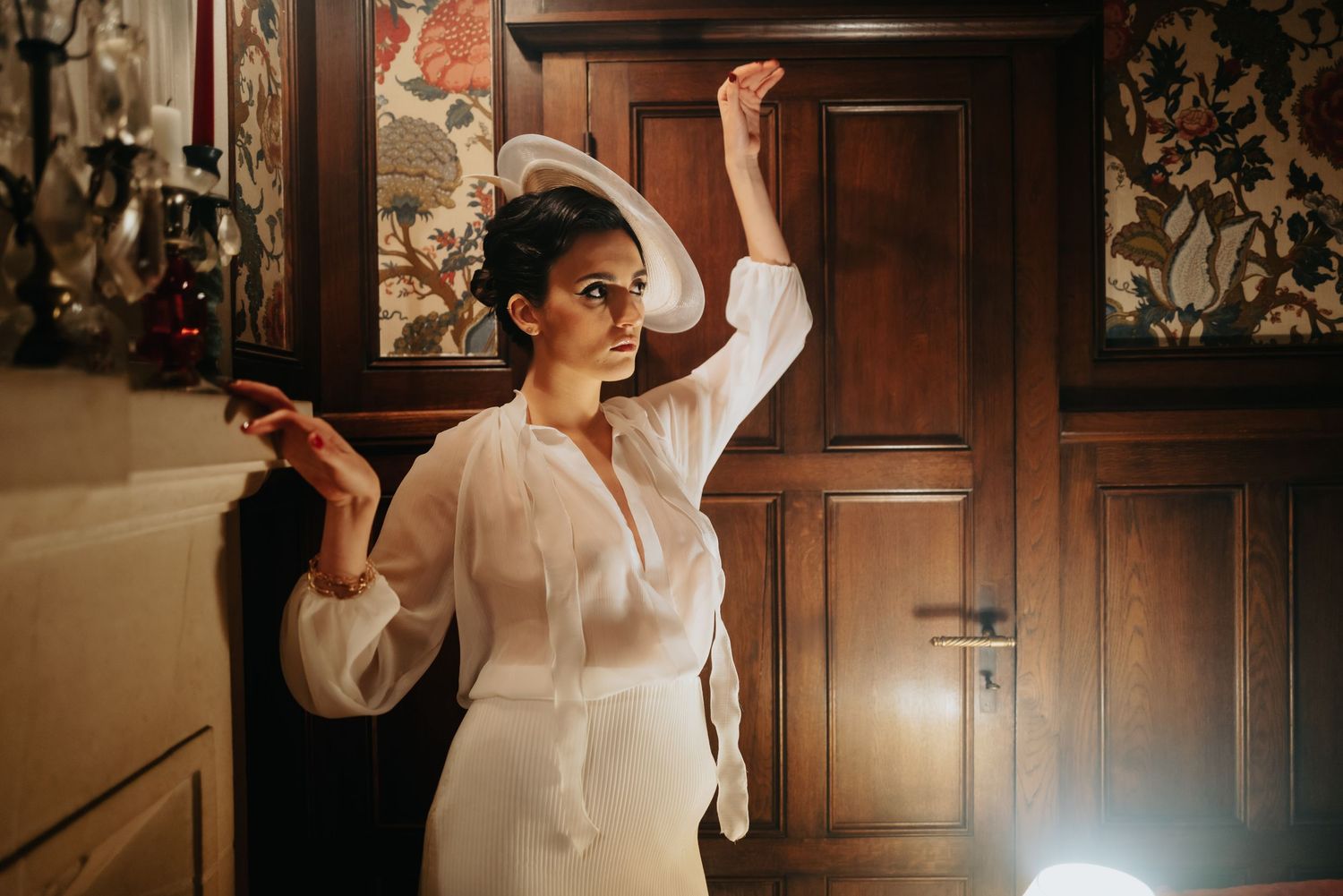 Woman in flowing white outfit strikes dramatic dance pose against wooden paneled wall.