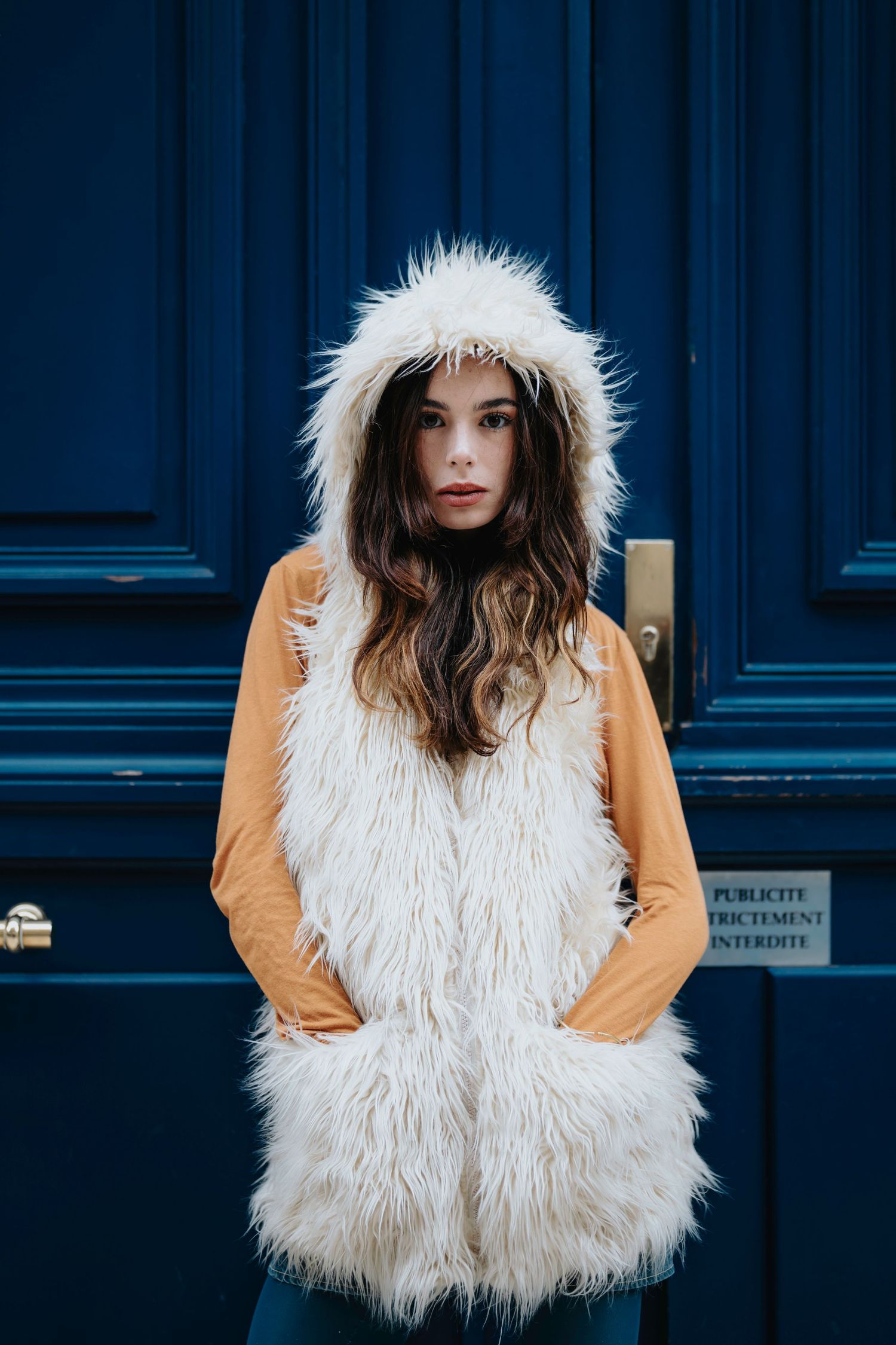 Street style portrait featuring white furry vest with hood against blue door background.