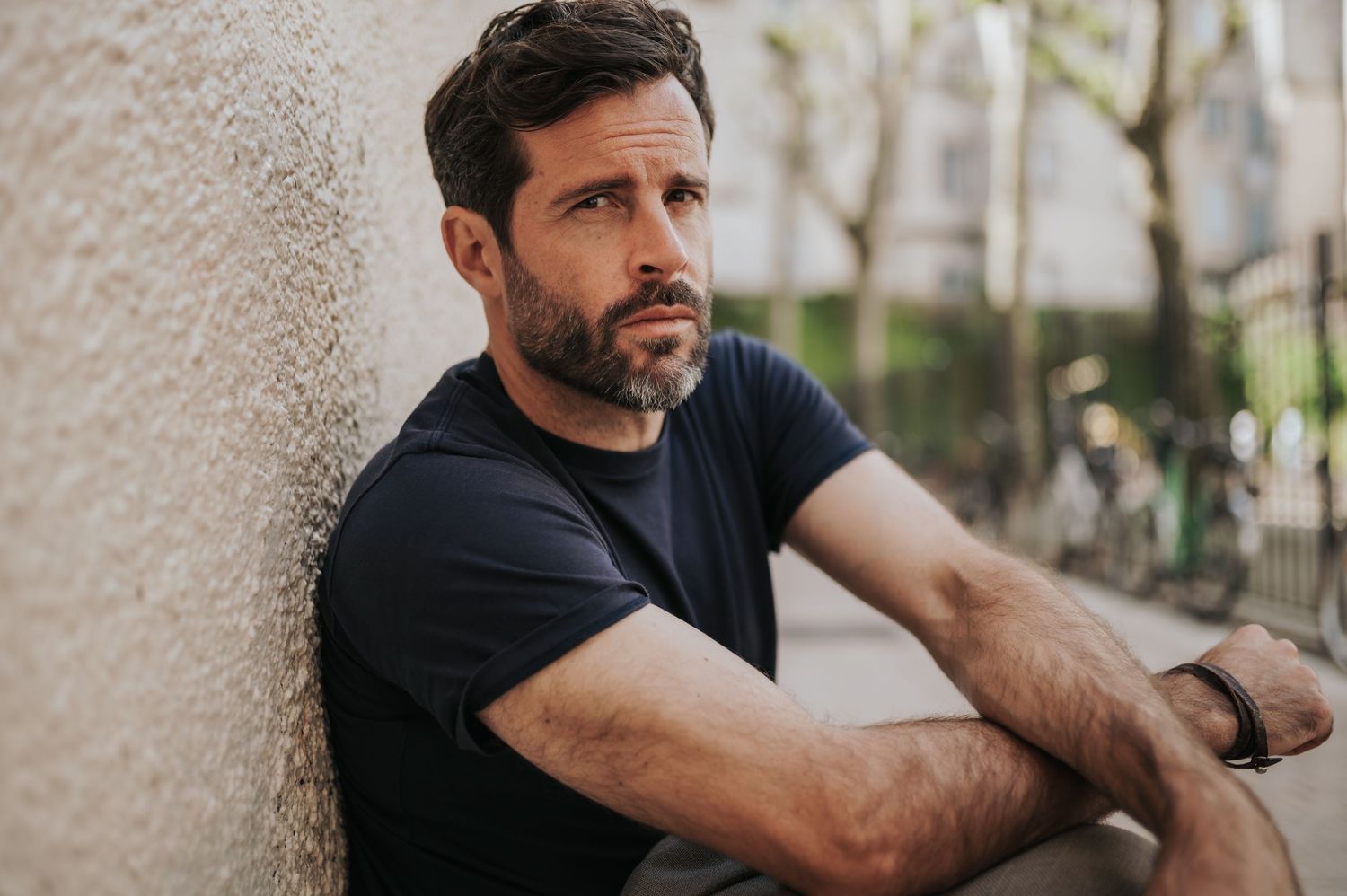 Casual portrait of man in navy t-shirt leaning against textured wall outdoors.