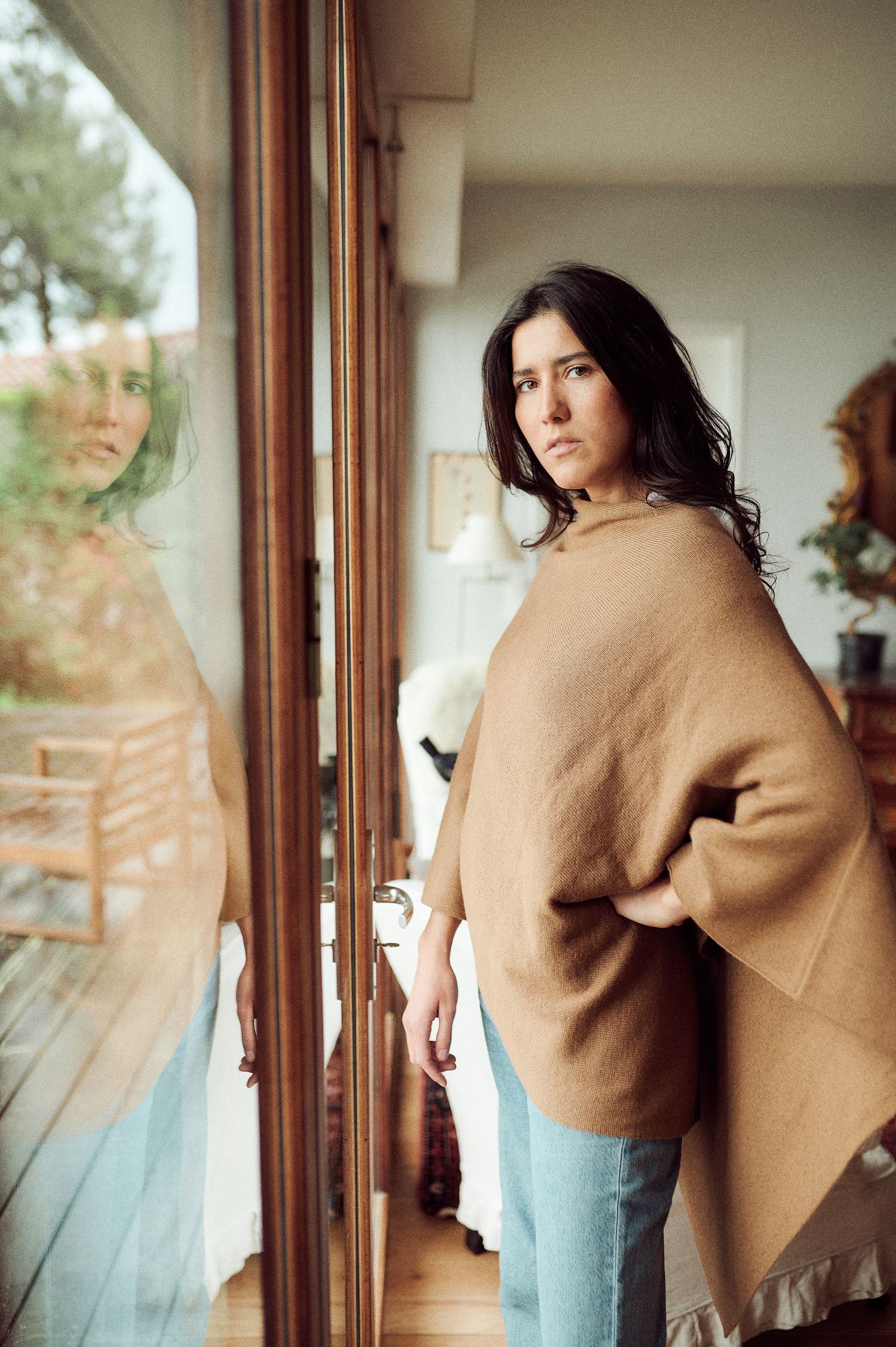 Model wearing camel colored wrap and denim poses against bright windows in modern interior.
