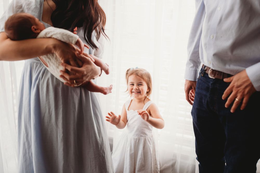 A child in a white dress joyfully claps while standing between two adults in a bright sunlit room.