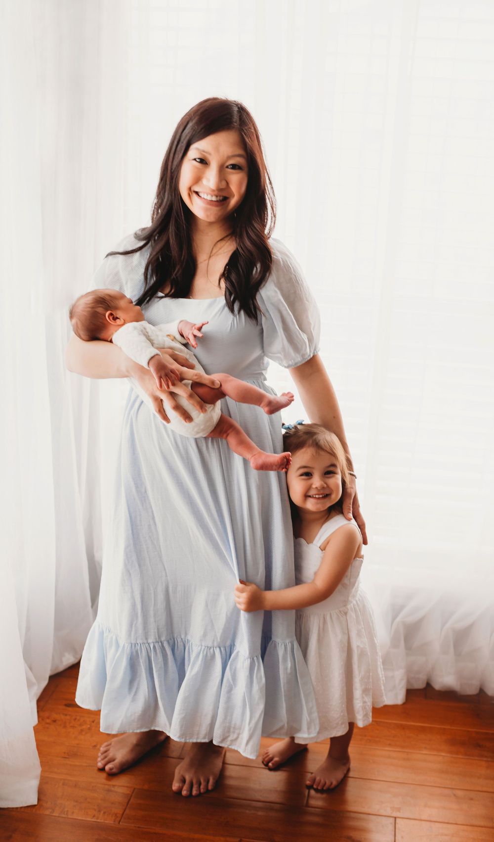 A woman in a light blue dress stands with children on hardwood floors by white curtains in a bright indoor setting.