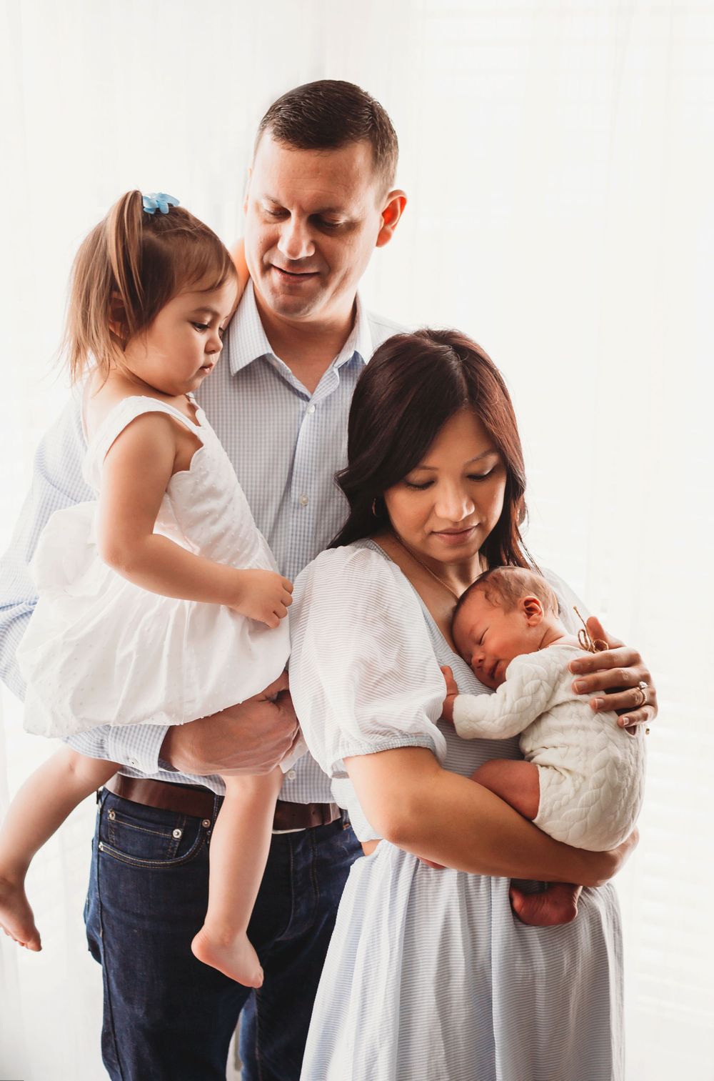 A family poses tenderly together, with a young child in a white dress and a newborn baby being held.