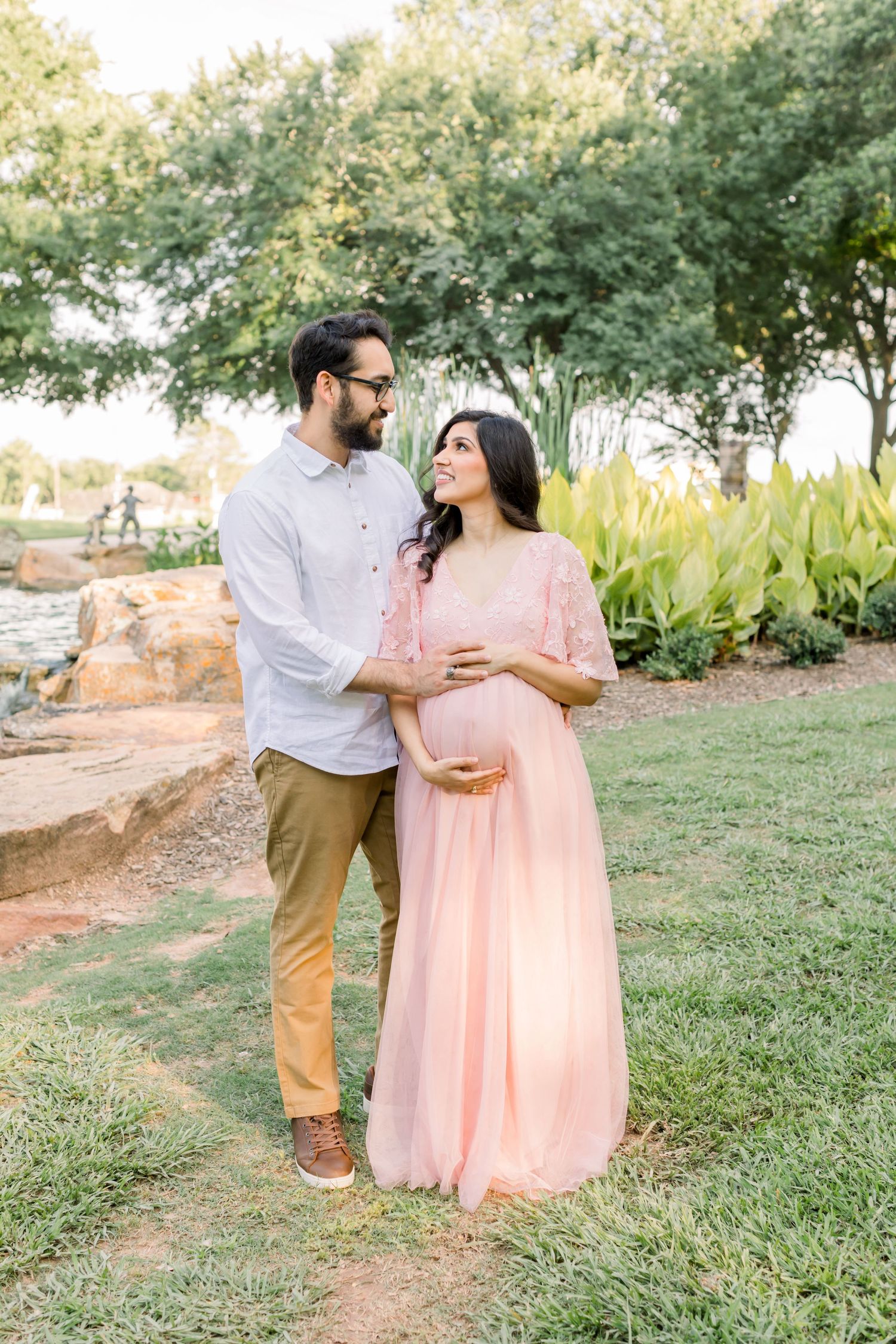 A couple shares a tender moment outdoors while standing on grass with trees in background.