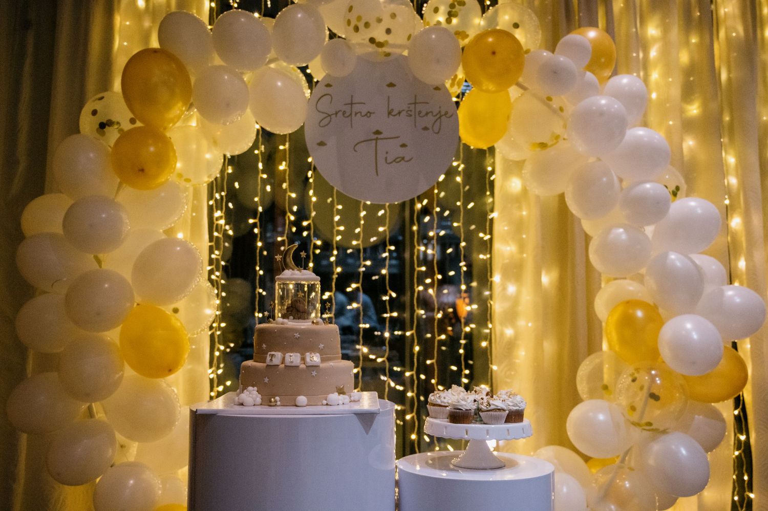 Elegant wedding cake display with white and gold balloon arch and fairy light backdrop.