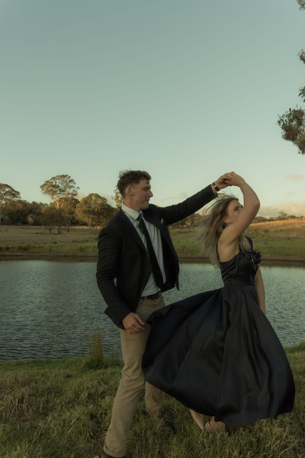 Couple in formal attire dancing together by a lake during golden hour.