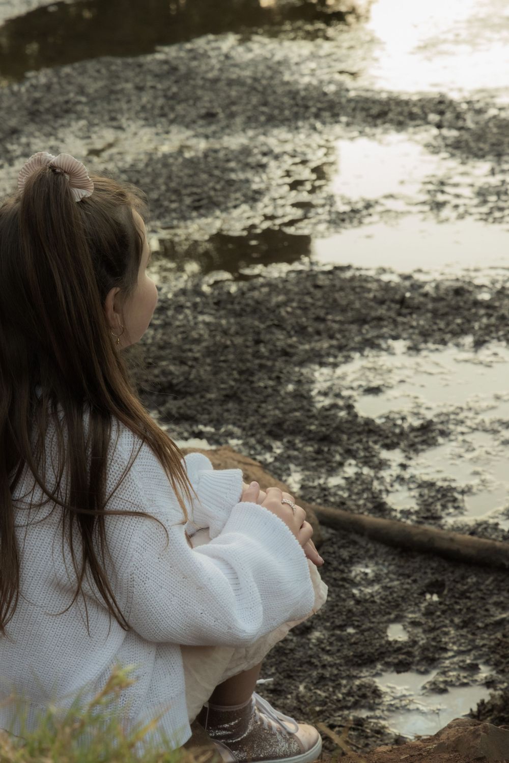 Child sitting quietly by a reflective pond wearing a white sweater in soft natural light.