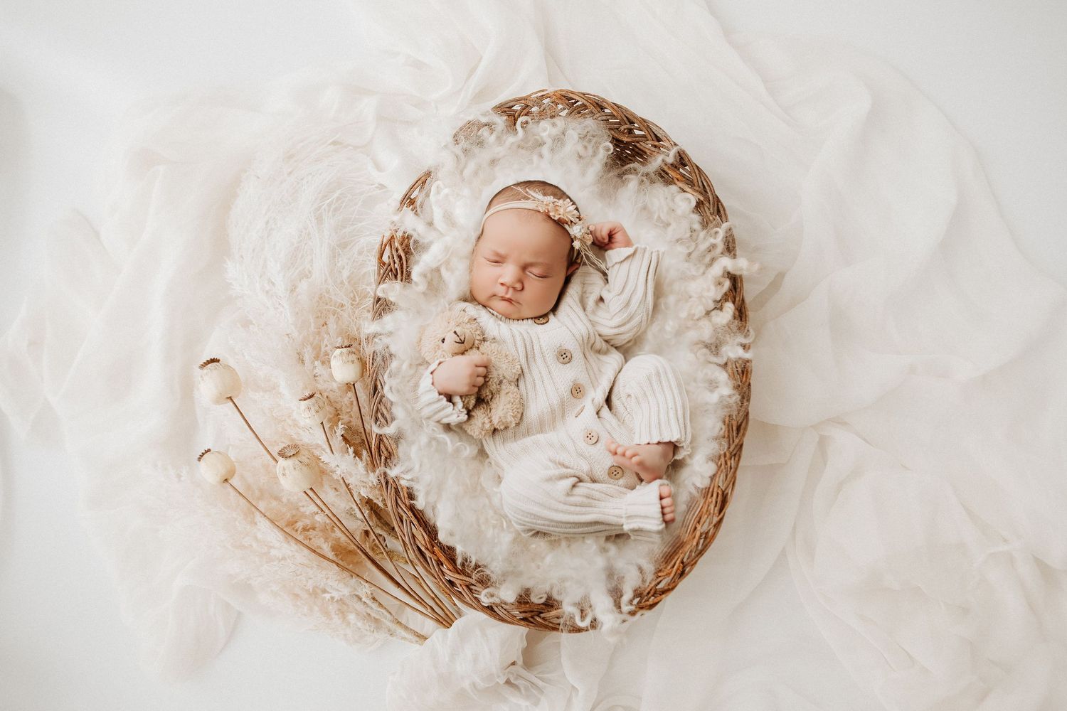 Sleeping newborn baby swaddled in white fabric nestled in a cozy cream-colored basket with dried flowers.