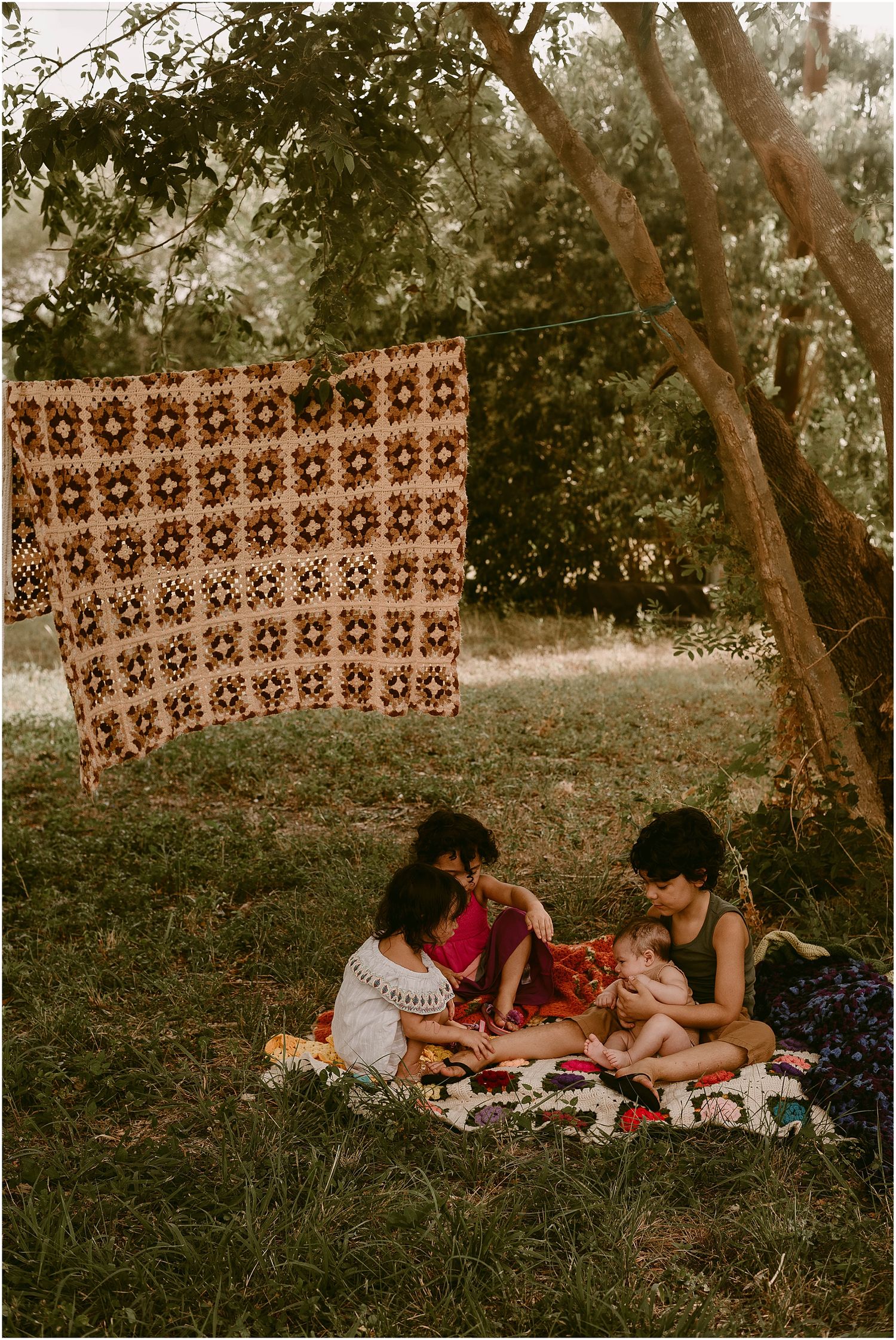 A family enjoys a picnic under trees with a brown patterned fabric hanging as a backdrop.