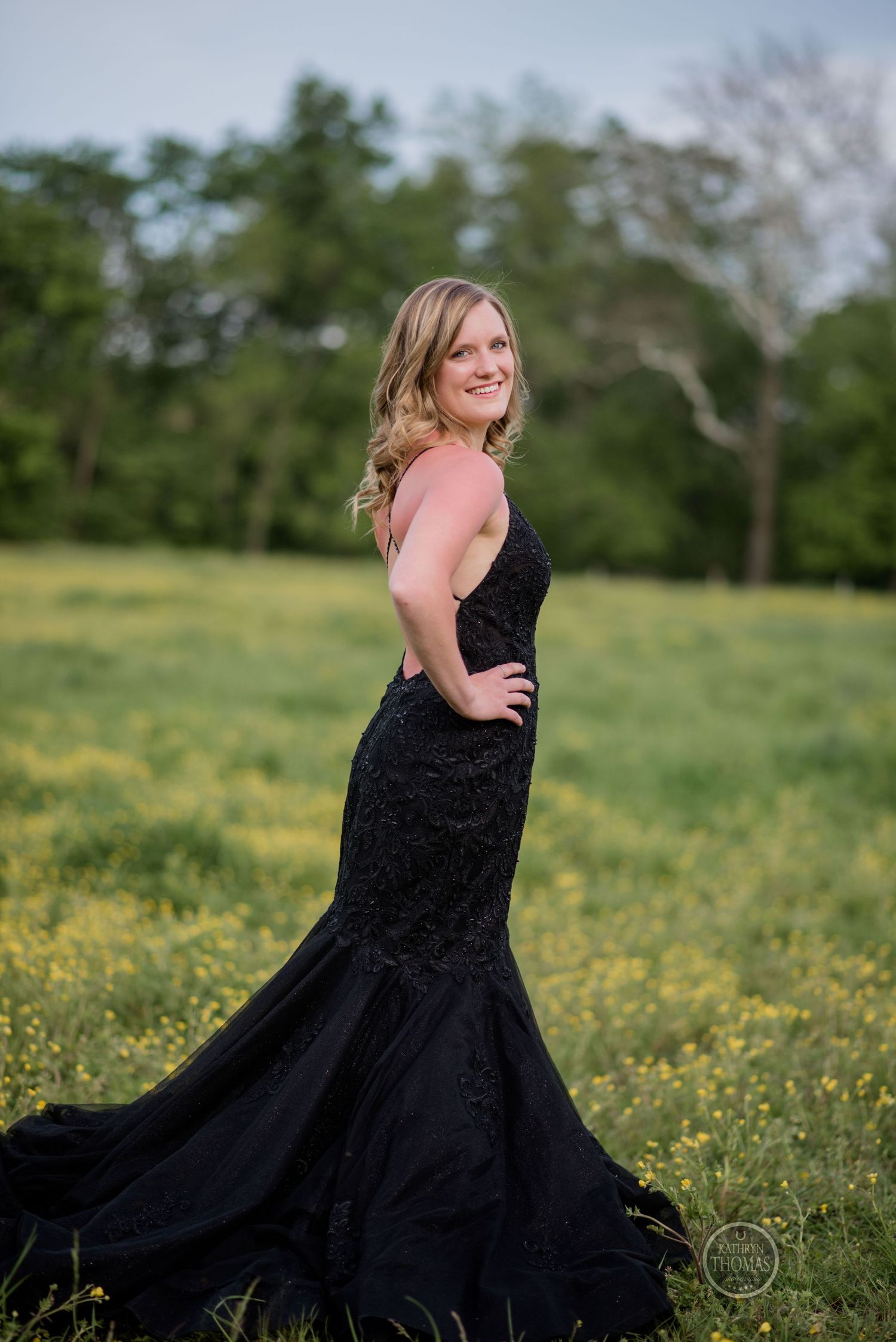 Woman wearing black mermaid gown poses in meadow filled with yellow spring flowers.
