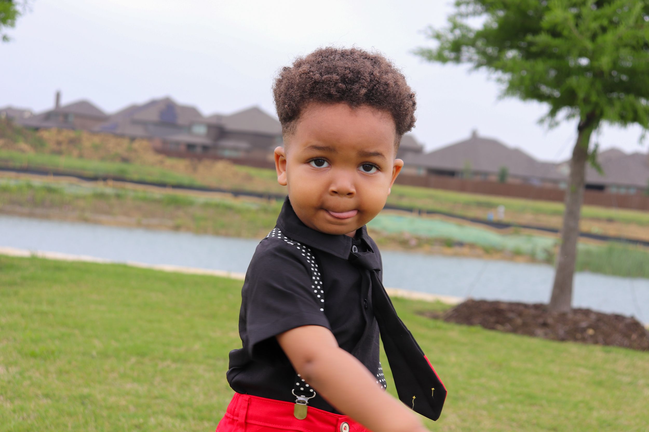 Small child in black shirt and red bottoms poses in grassy area with houses in background.