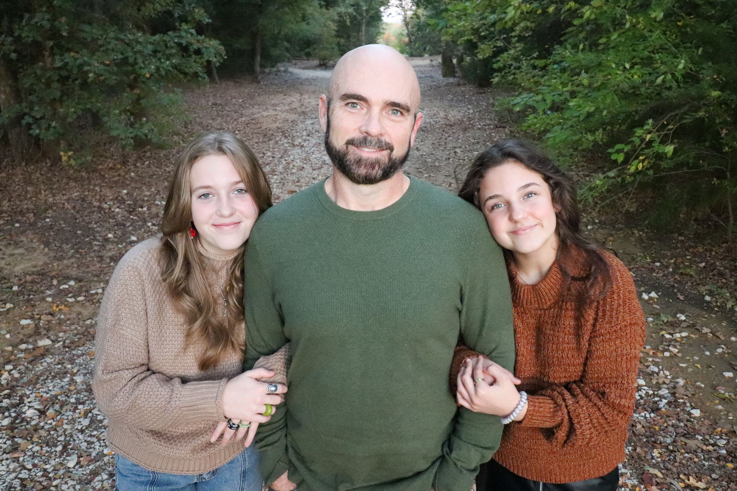 Three people in casual sweaters pose together on wooded path during outdoor portrait session.