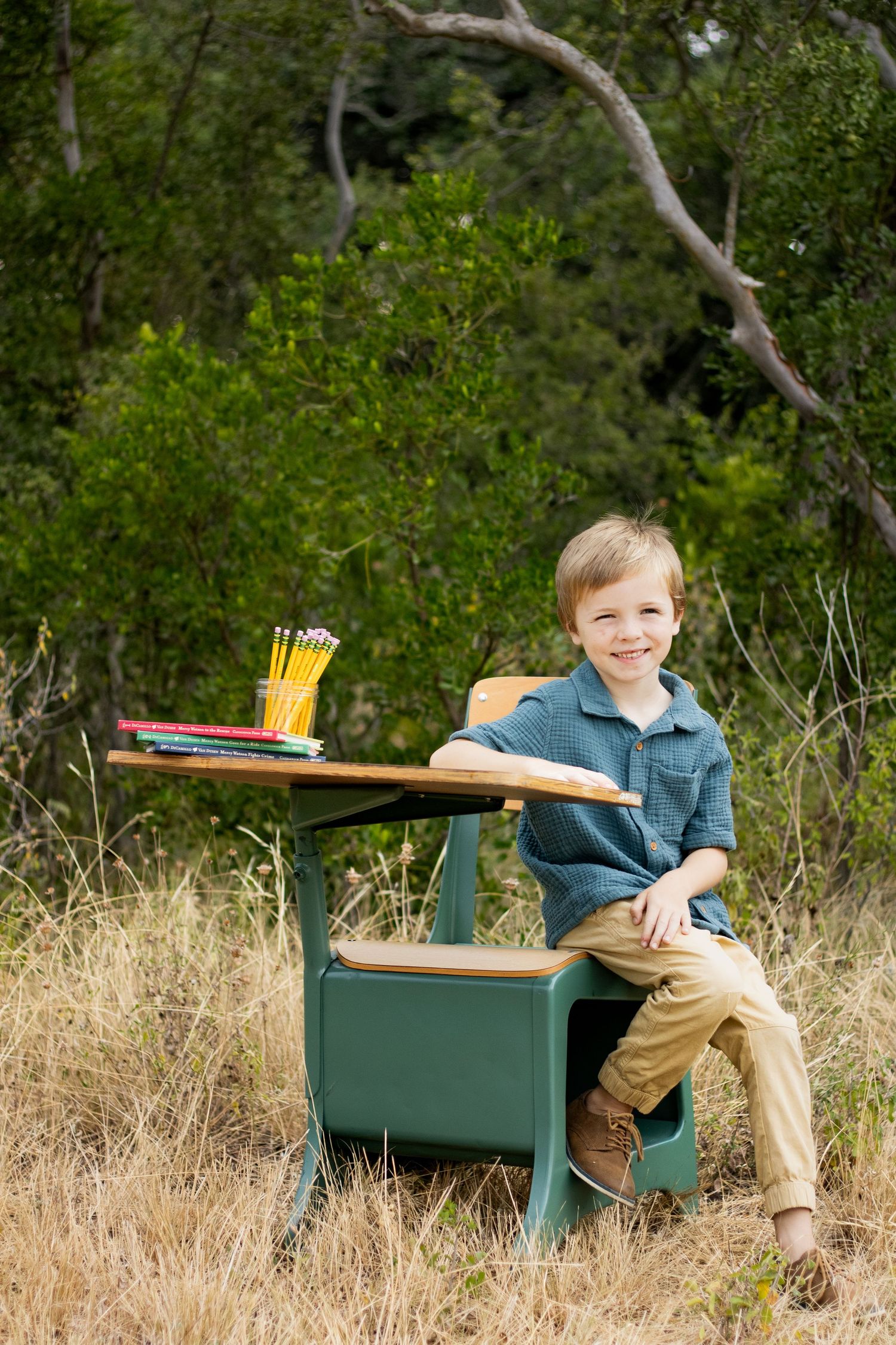 Young boy in casual clothing sitting on green vintage school desk in outdoor field setting.