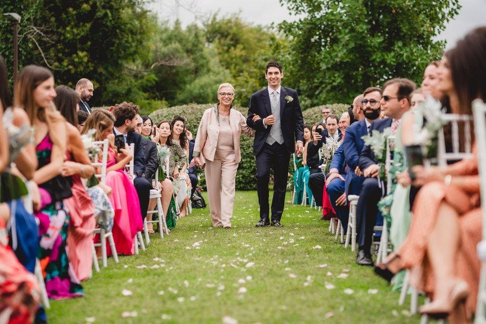Outdoor wedding ceremony with guests seated on both sides of a flower-lined aisle.
