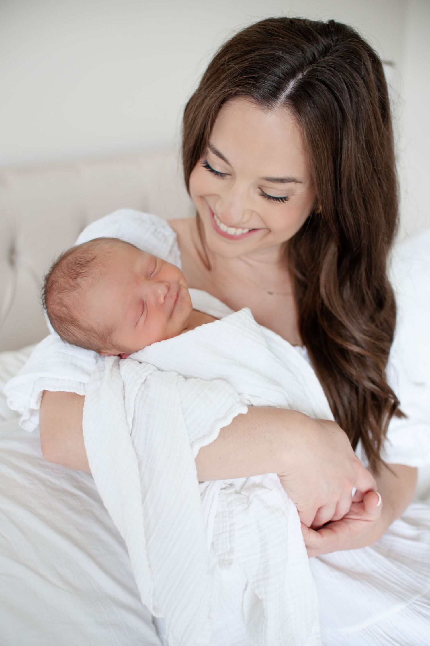 mother in all white smiles down on baby wrapped in white swaddle. image from an at-home newborn session