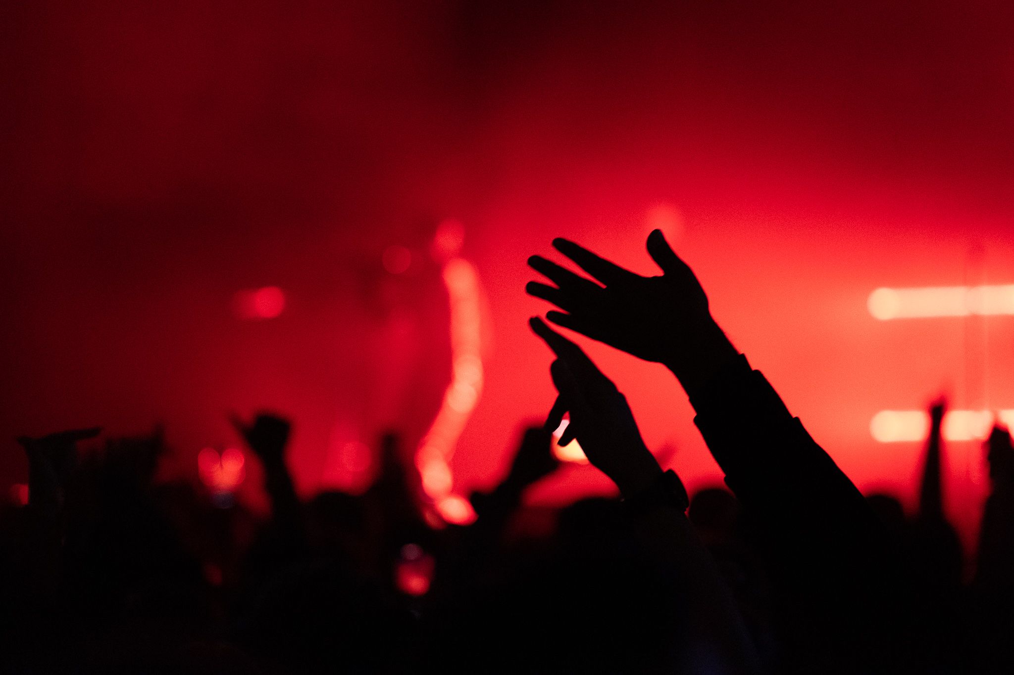 Silhouetted crowd hands raised at concert with red atmospheric lighting.