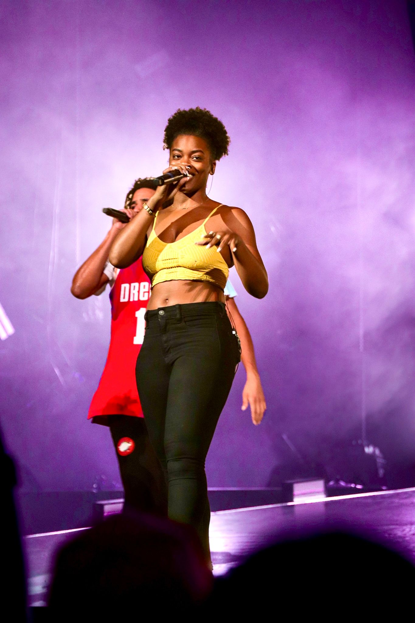 Performer in yellow crop top energetically singing on stage with purple lighting effects.