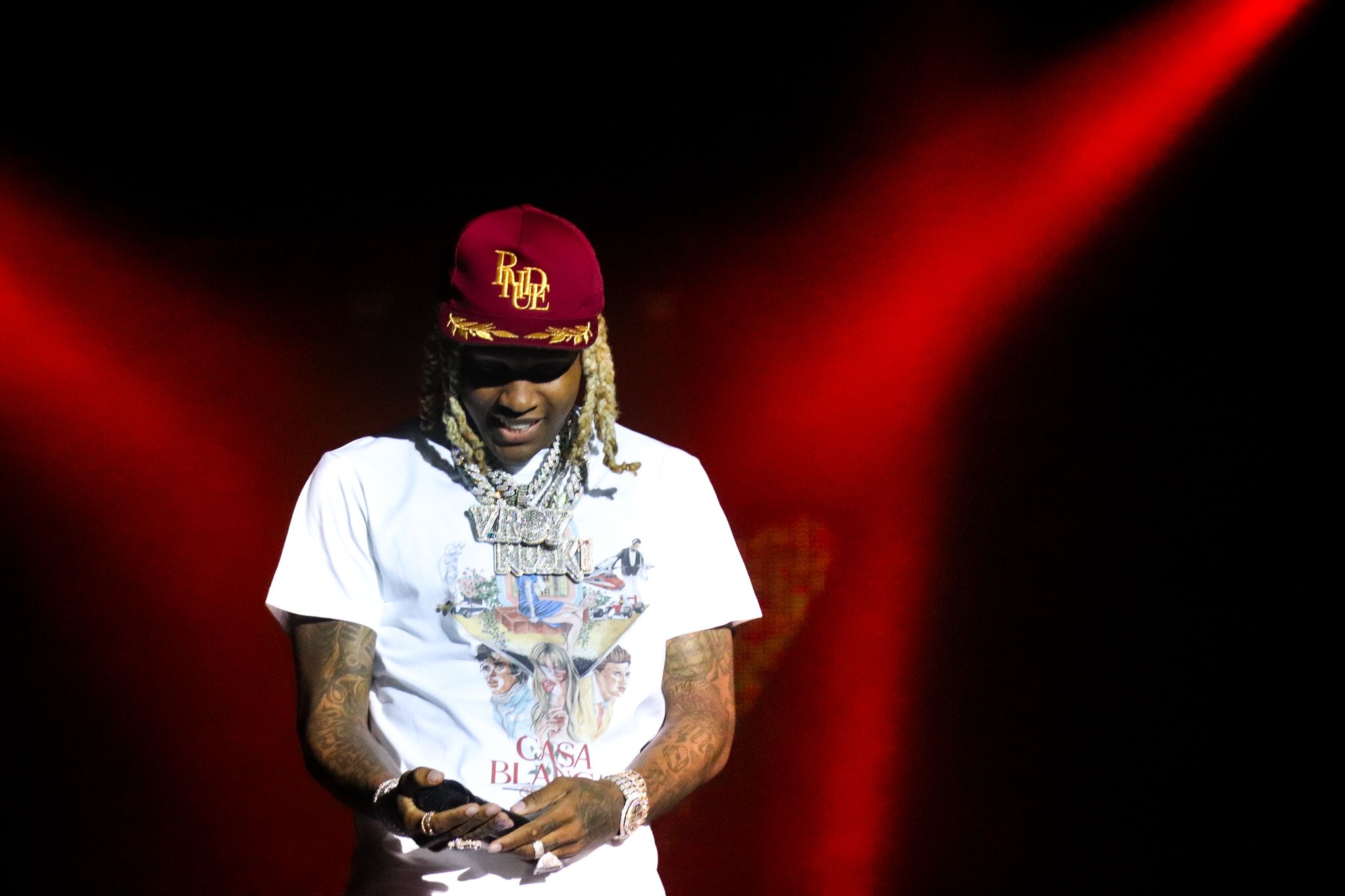 Artist in white t-shirt and burgundy cap performs under dramatic red stage lighting.