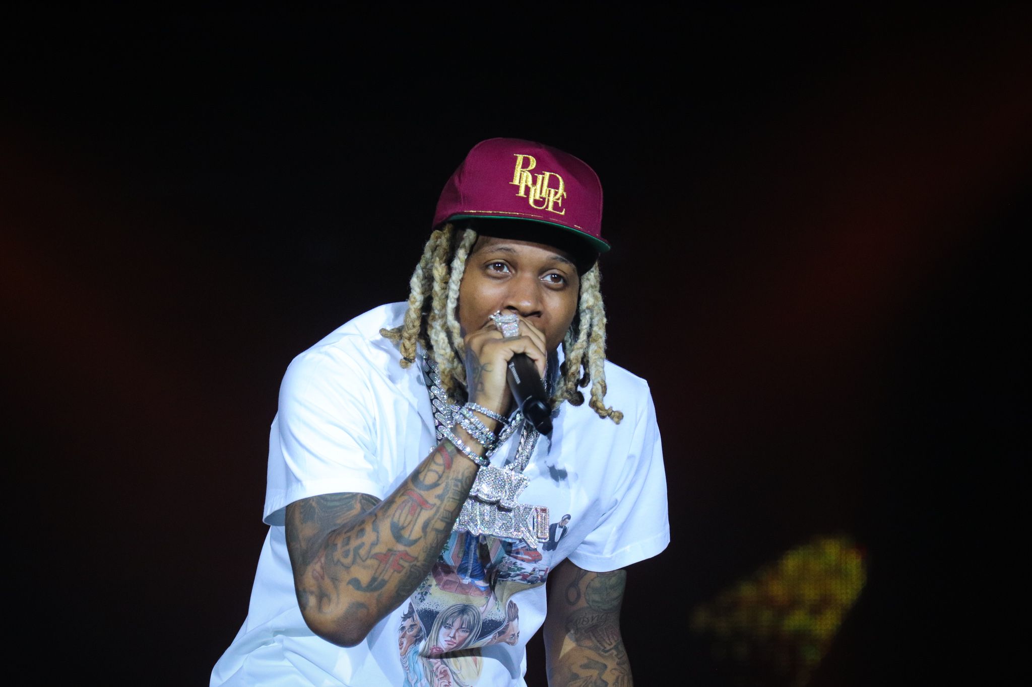 Performer in white t-shirt and burgundy cap singing into microphone on dark stage.