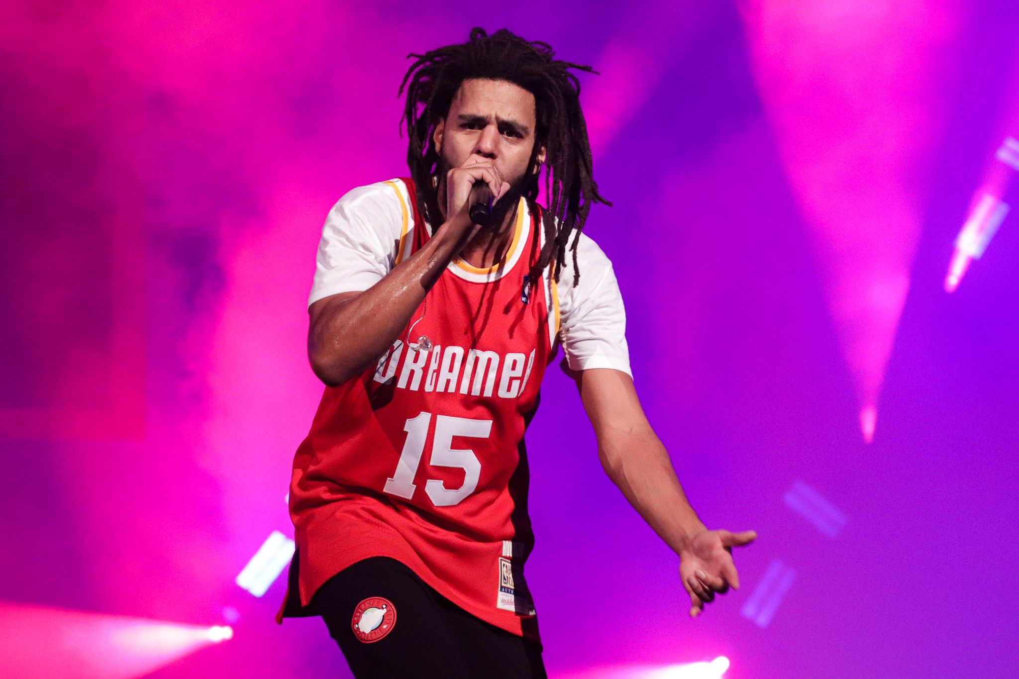 Energetic performer in red basketball jersey singing passionately on stage with purple lighting.