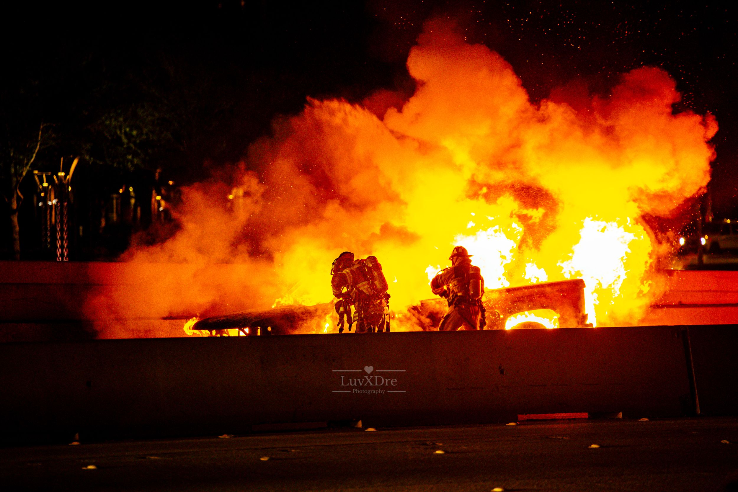 Silhouetted figures sit in front of large flames and smoke at night during an intense fire scene.