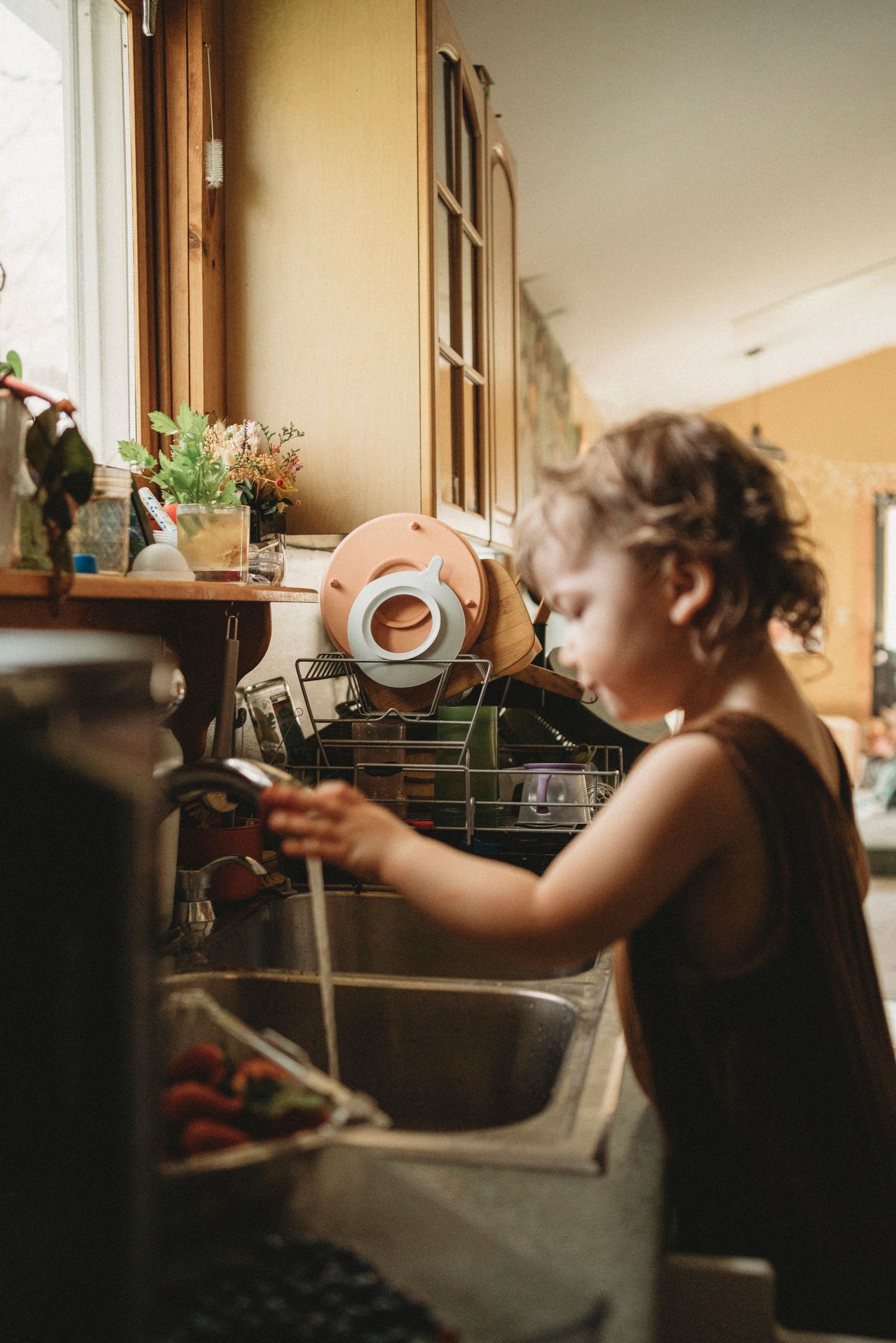 Child helping with dishes at kitchen sink in warm natural lighting.