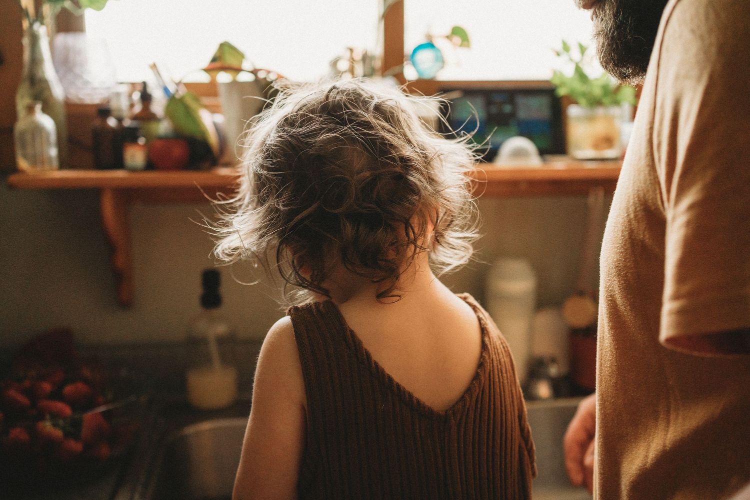 Person with curly hair wearing a brown tank top stands at a kitchen counter in warm lighting.