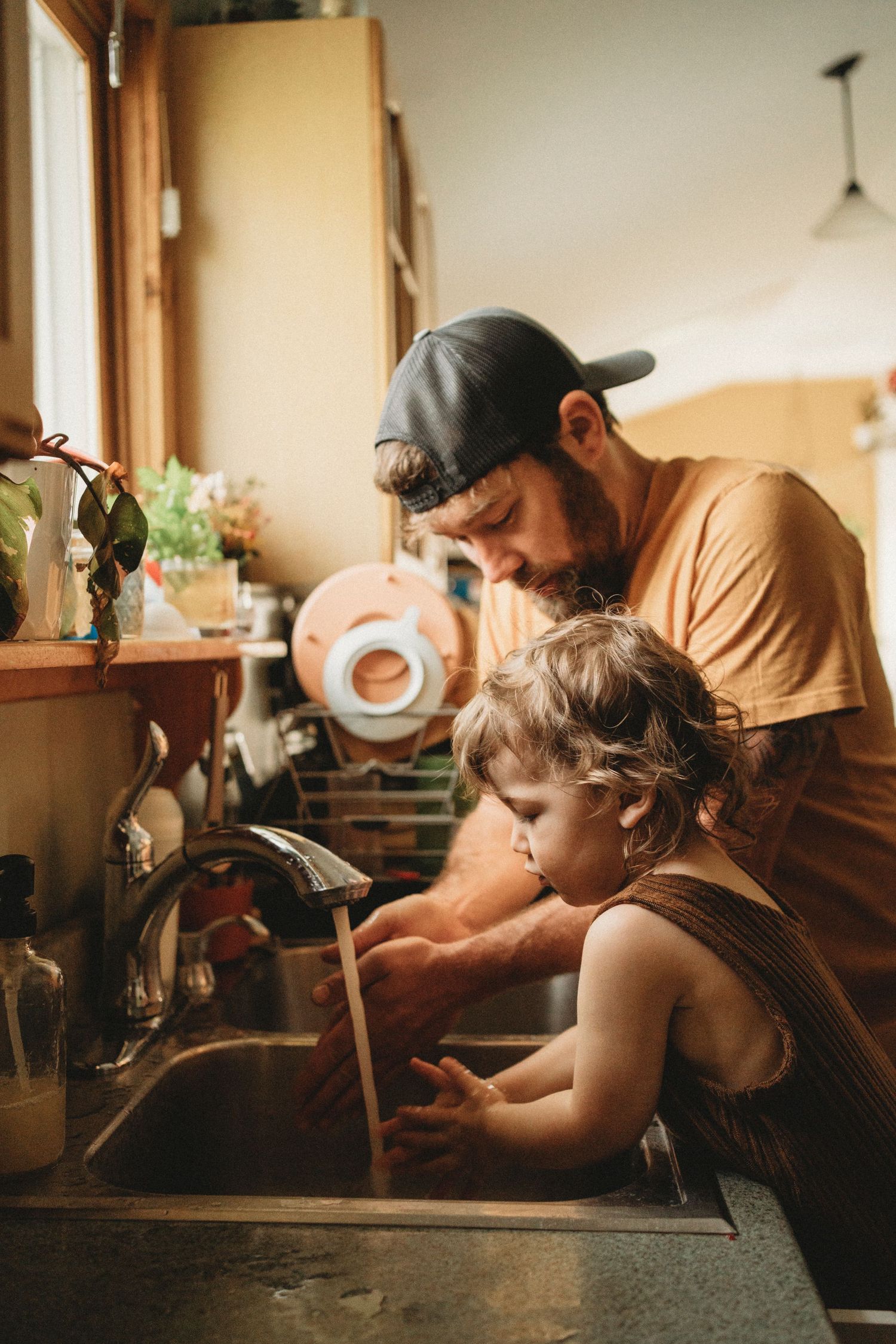 Father and young child doing dishes together at a kitchen sink with warm sunlight streaming in.