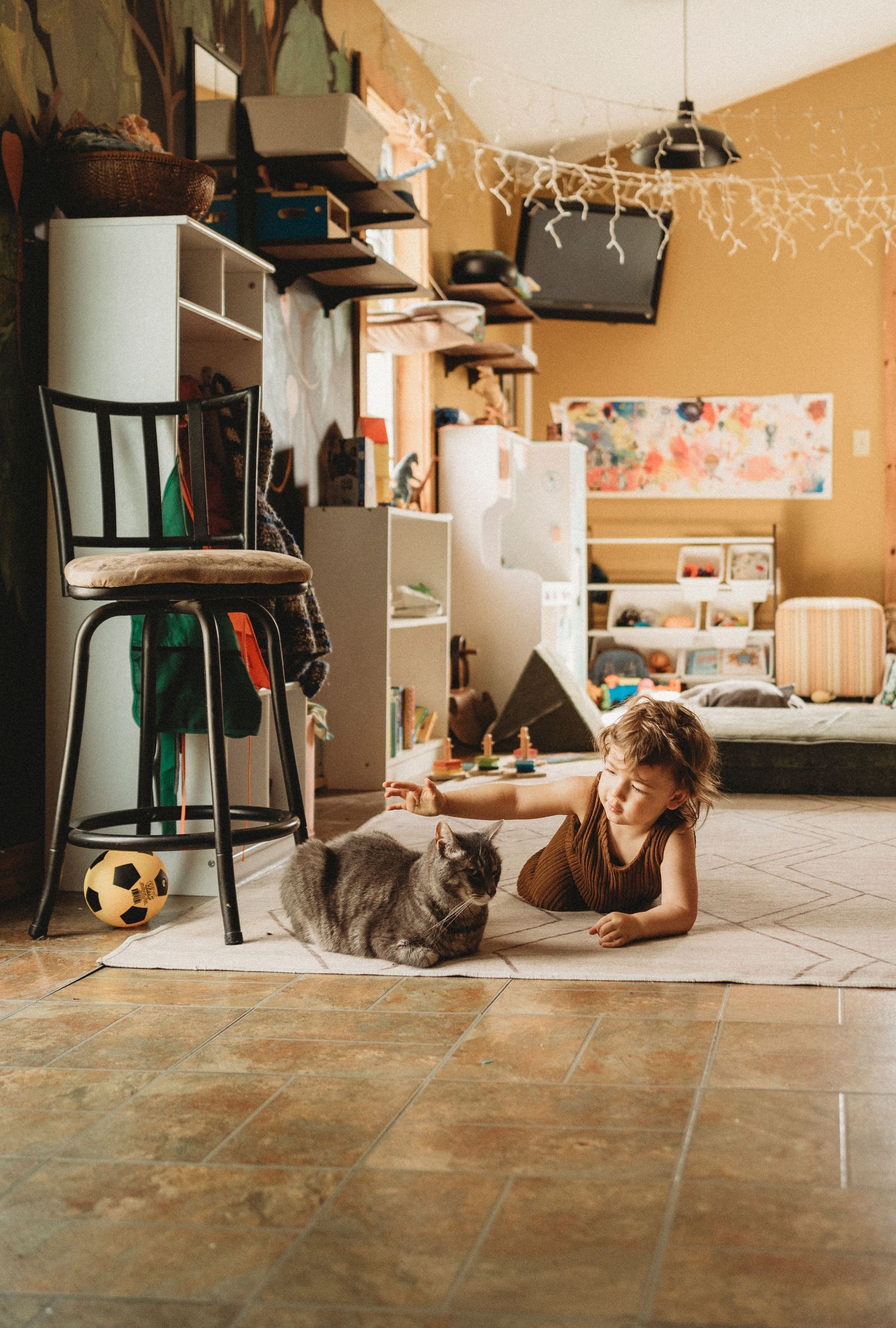 Child lounging on the floor with a grey cat in a cozy room with string lights and storage shelves.