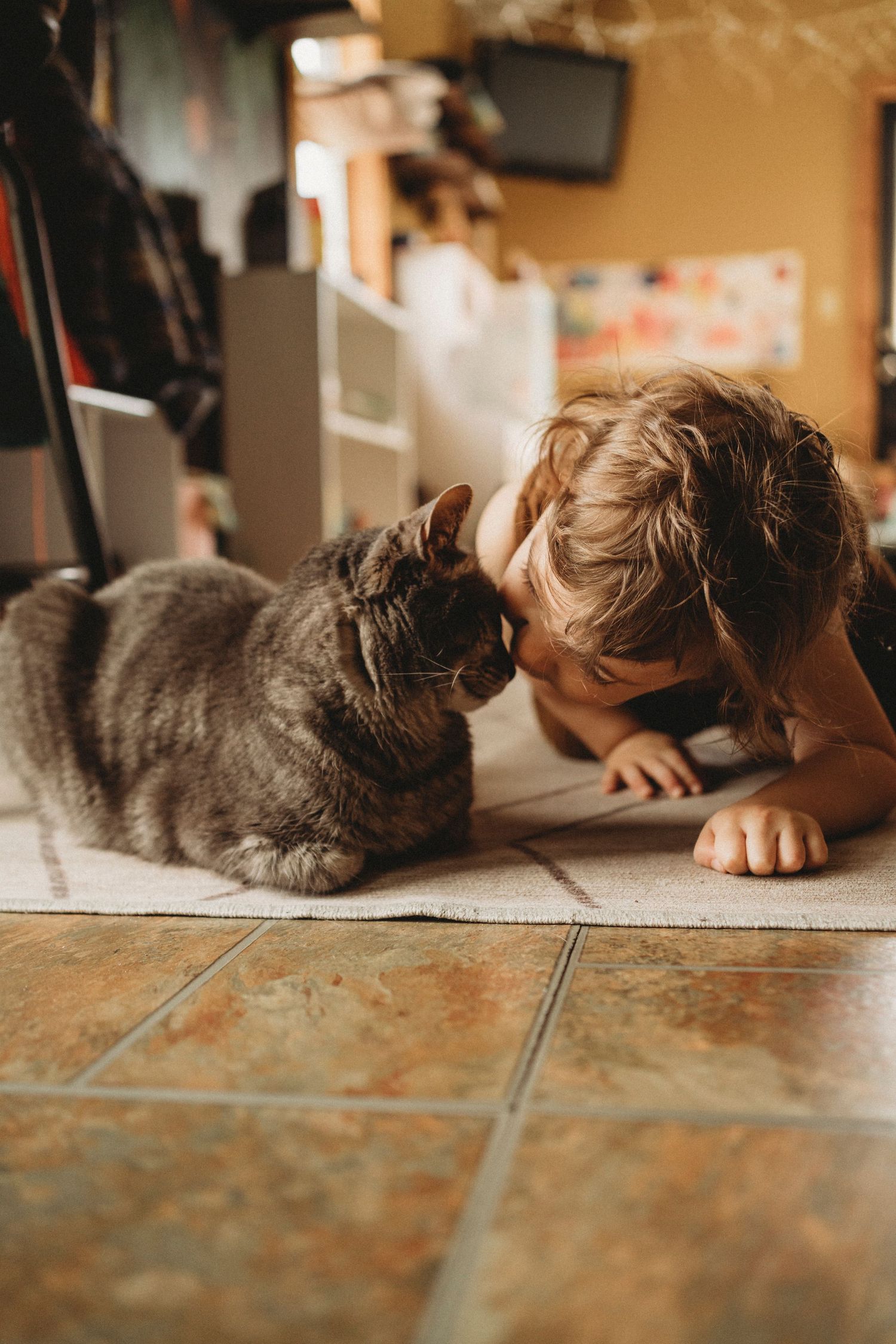 Child laying on tile floor nose-to-nose with a grey cat in an intimate moment.