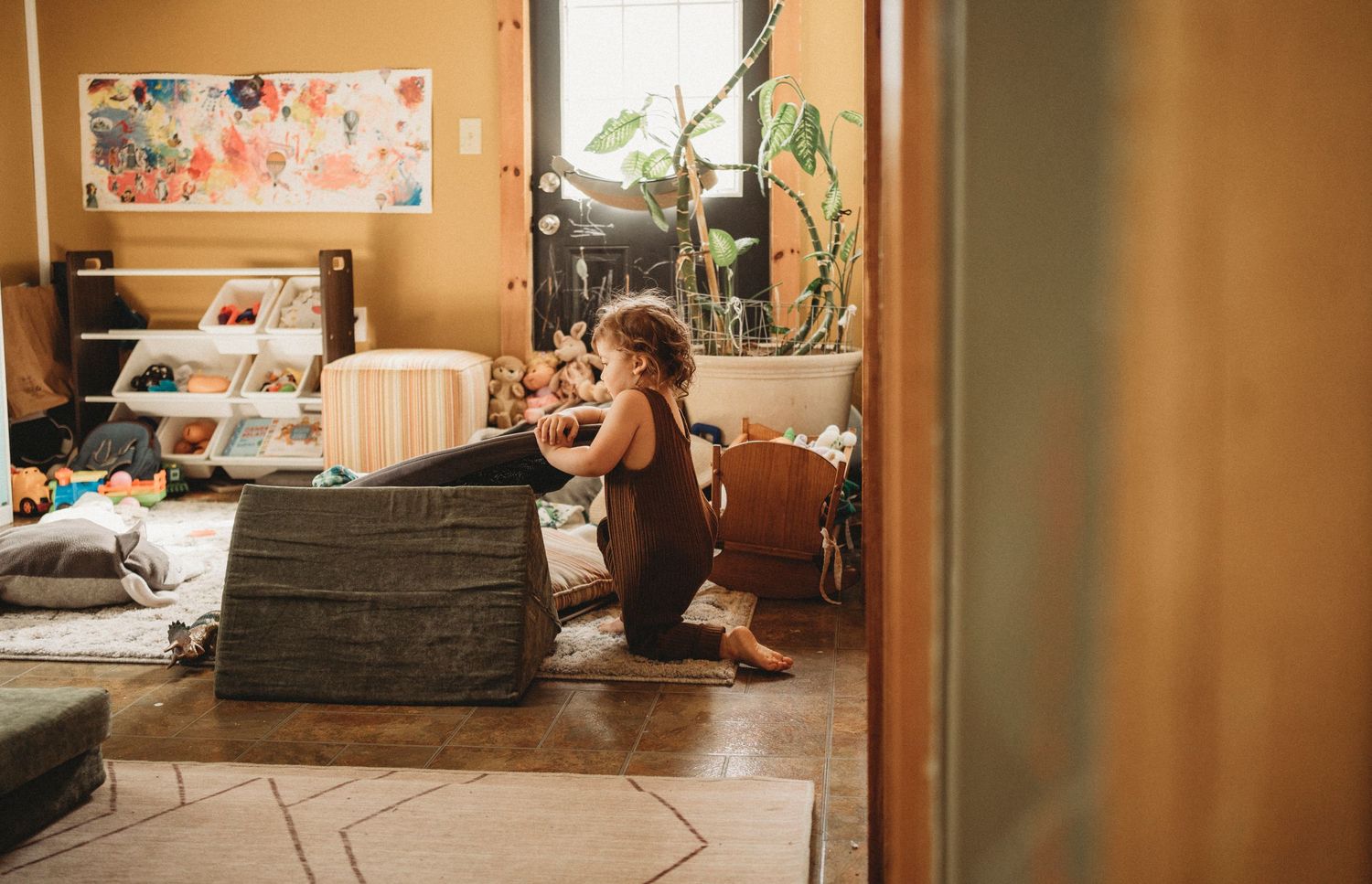 View through doorway of child sitting on floor near storage furniture in a warmly lit room.