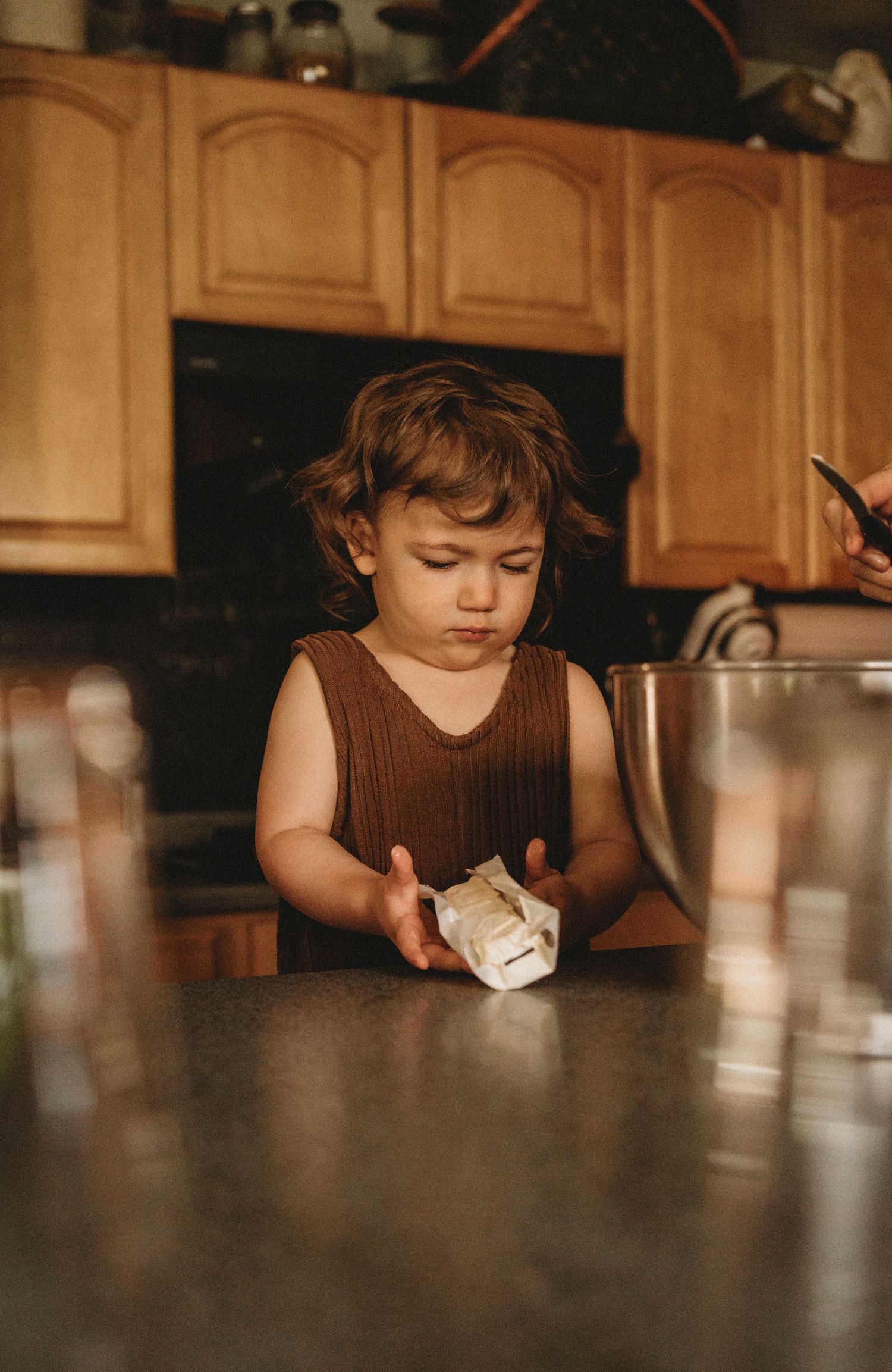 Small child in brown tank top concentrating while handling something at wooden kitchen table.