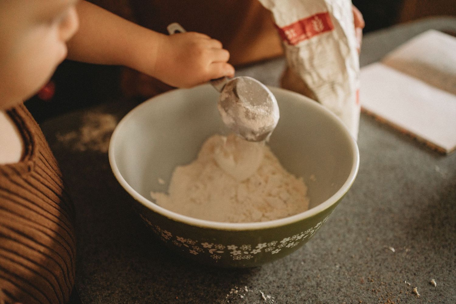 A small hand pours dry ingredients into a large green and white mixing bowl.