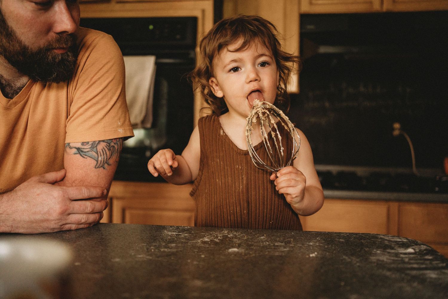 A young child licks a baking utensil while cooking in the kitchen.