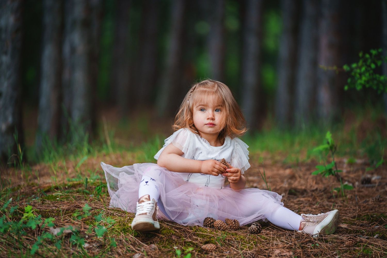 A small child in a pink tulle dress sits on a forest path surrounded by tall pine trees.