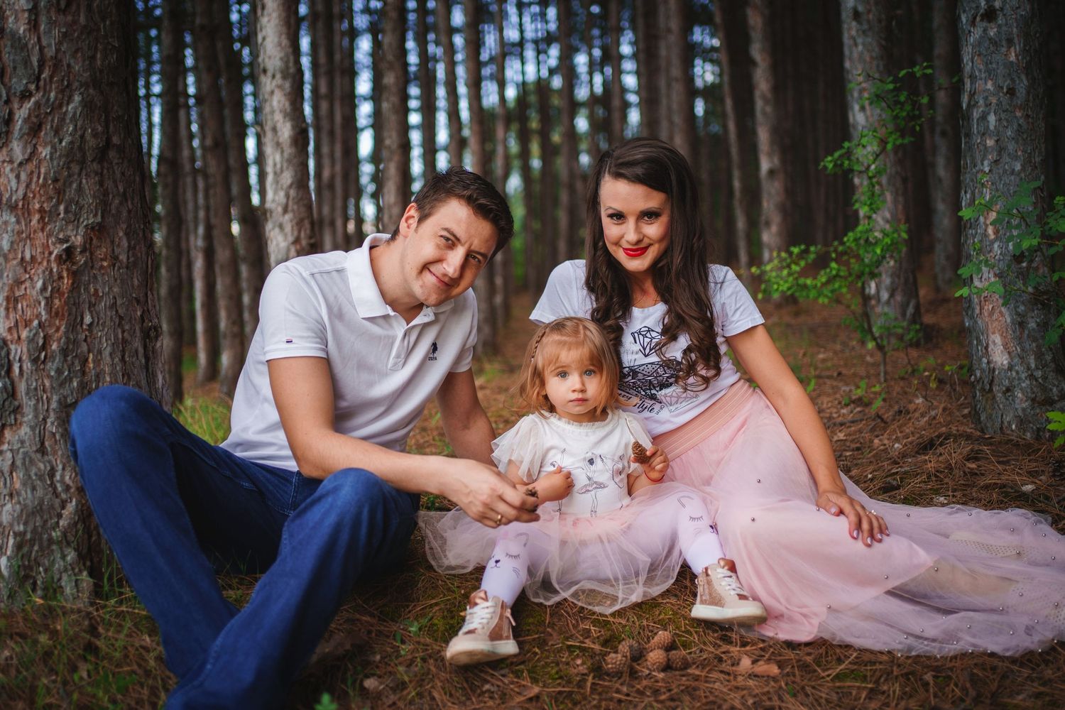 Family sitting together on forest floor surrounded by tall pine trees.
