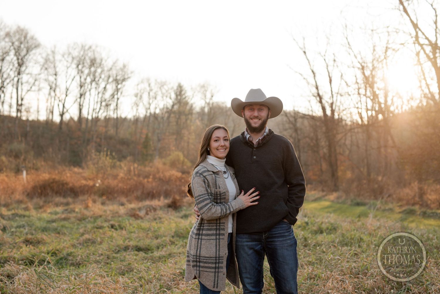 A romantic couple poses together in a field at sunset wearing autumn attire with trees in background.
