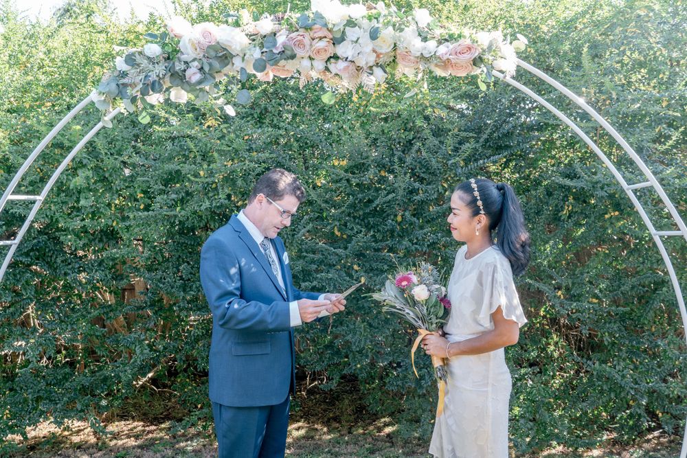 groom in blue suit reading vows to asian bride in white dress and holding bouquet under decorated arbour