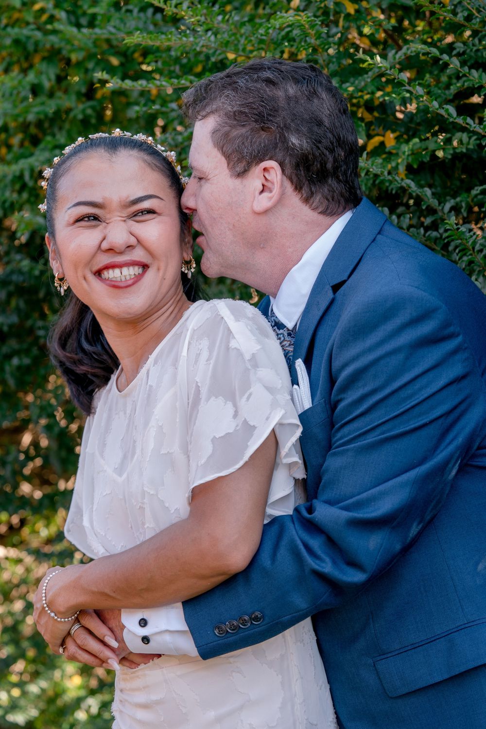 mature asian bride smiling as mature groom in blue suit whispers in her ear