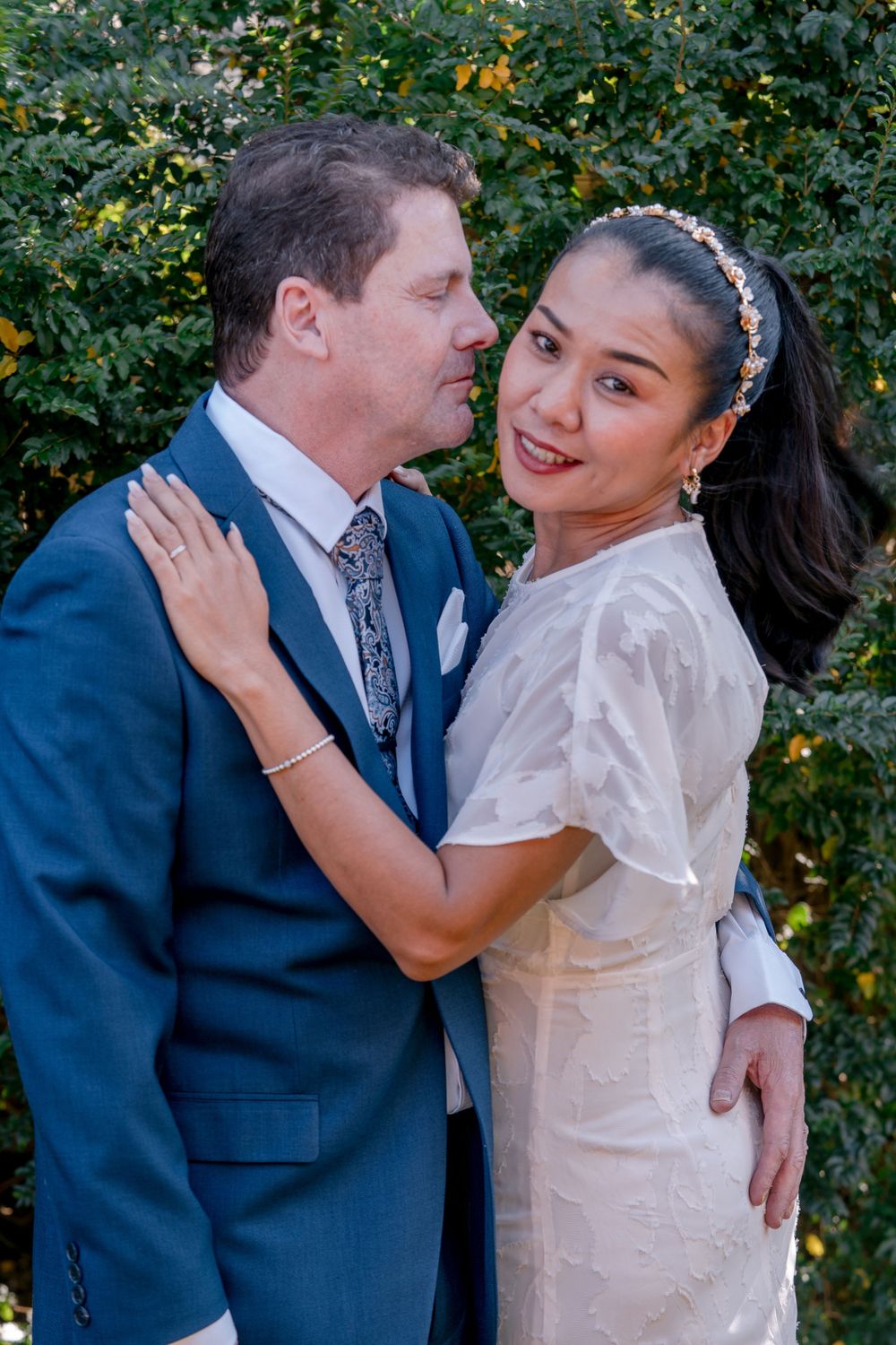 mature groom in blue suit posing with mature asian bride in headband and slicked back hair