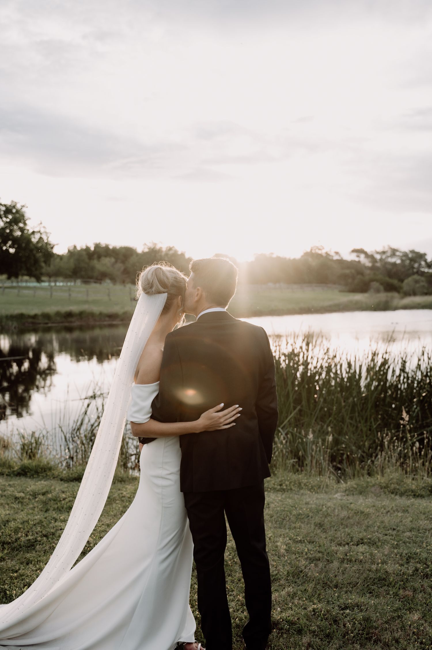 A couple in wedding attire embraces by a tranquil lake at sunset with a long veil flowing behind.